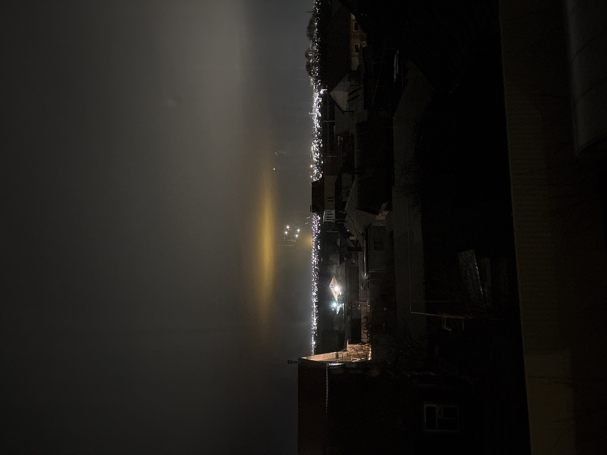 The image depicts a night scene of Ulyanovsk, Russia, taken from an elevated position. The view includes a river or large body of water on the left side of the image, obscured by heavy fog, and the illuminated cityscape along the waterfront on the right. The city lights create a vertical line of illumination that cuts through the fog. The foreground consists of darkened building structures, which suggest that the image was taken from a building overlooking the city.