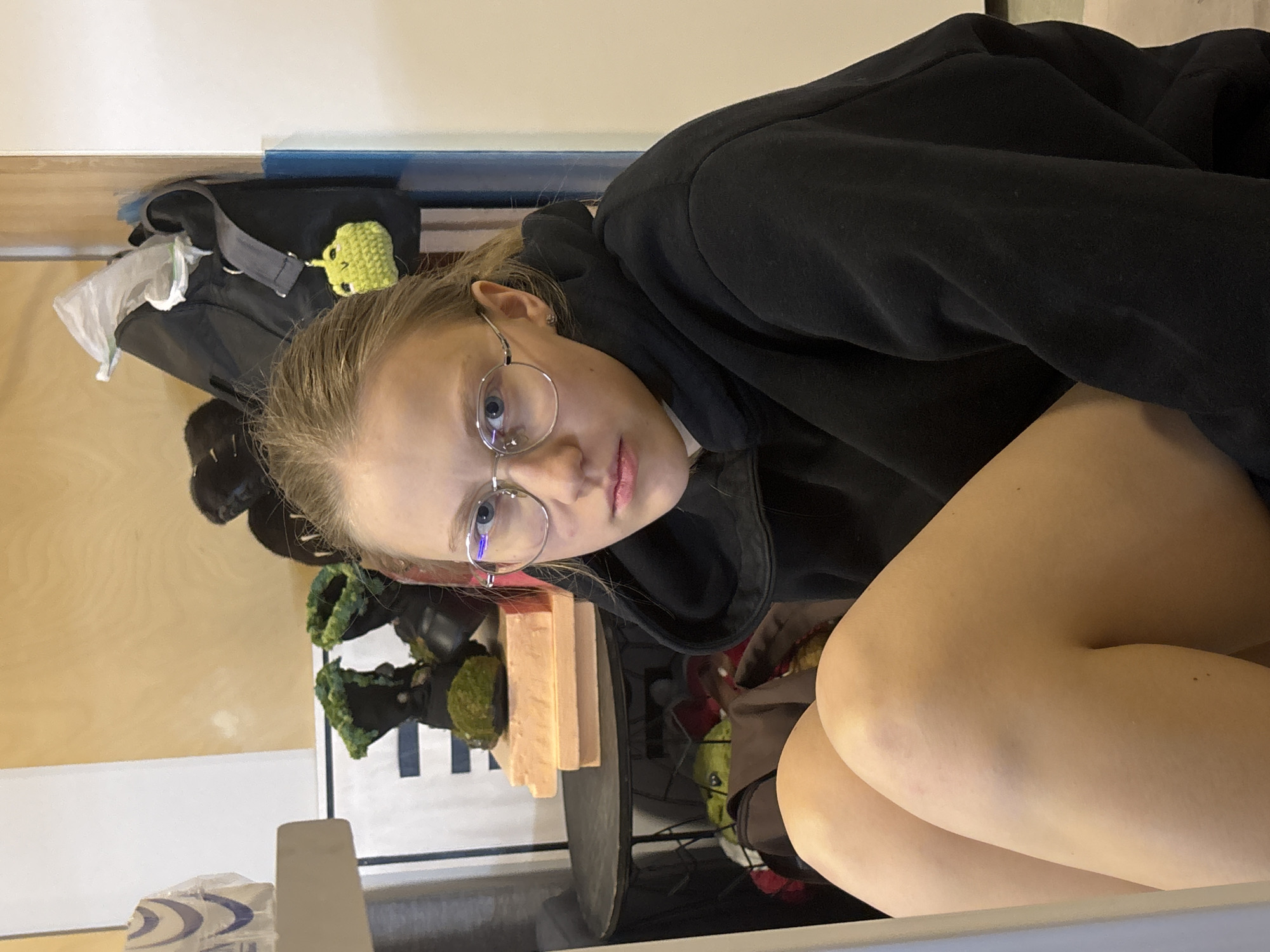A young woman with blond hair pulled back is wearing round glasses and a black hoodie, looking directly at the camera. She is sitting with her legs crossed. Behind her, on a shelf, is a black backpack with a green knitted frog attached. The background includes a wall with a blue stripe and various items on the shelf, including what appears to be plants and other miscellaneous objects. The location is likely indoors, possibly a room in a building in Perm, Russia, given the presence of the Russian language in the image data.