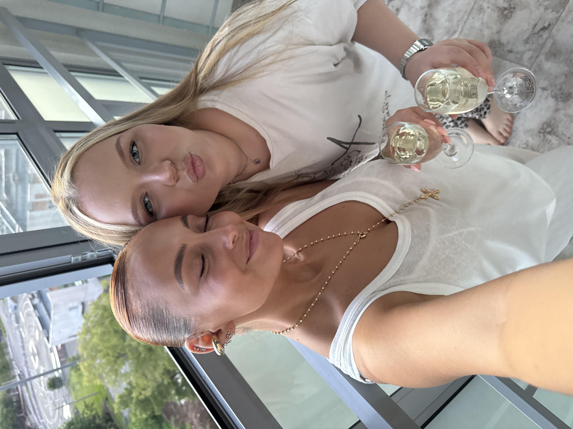 Two women are posing for a selfie. Both are wearing white t-shirts and holding champagne glasses. The blonde woman is pouting and has a watch on her wrist. The other woman is wearing gold jewelry including a necklace with a cross. A window is visible, reflecting the outdoors. Based on the reflection, the location may be Izhevsk, Russia.
