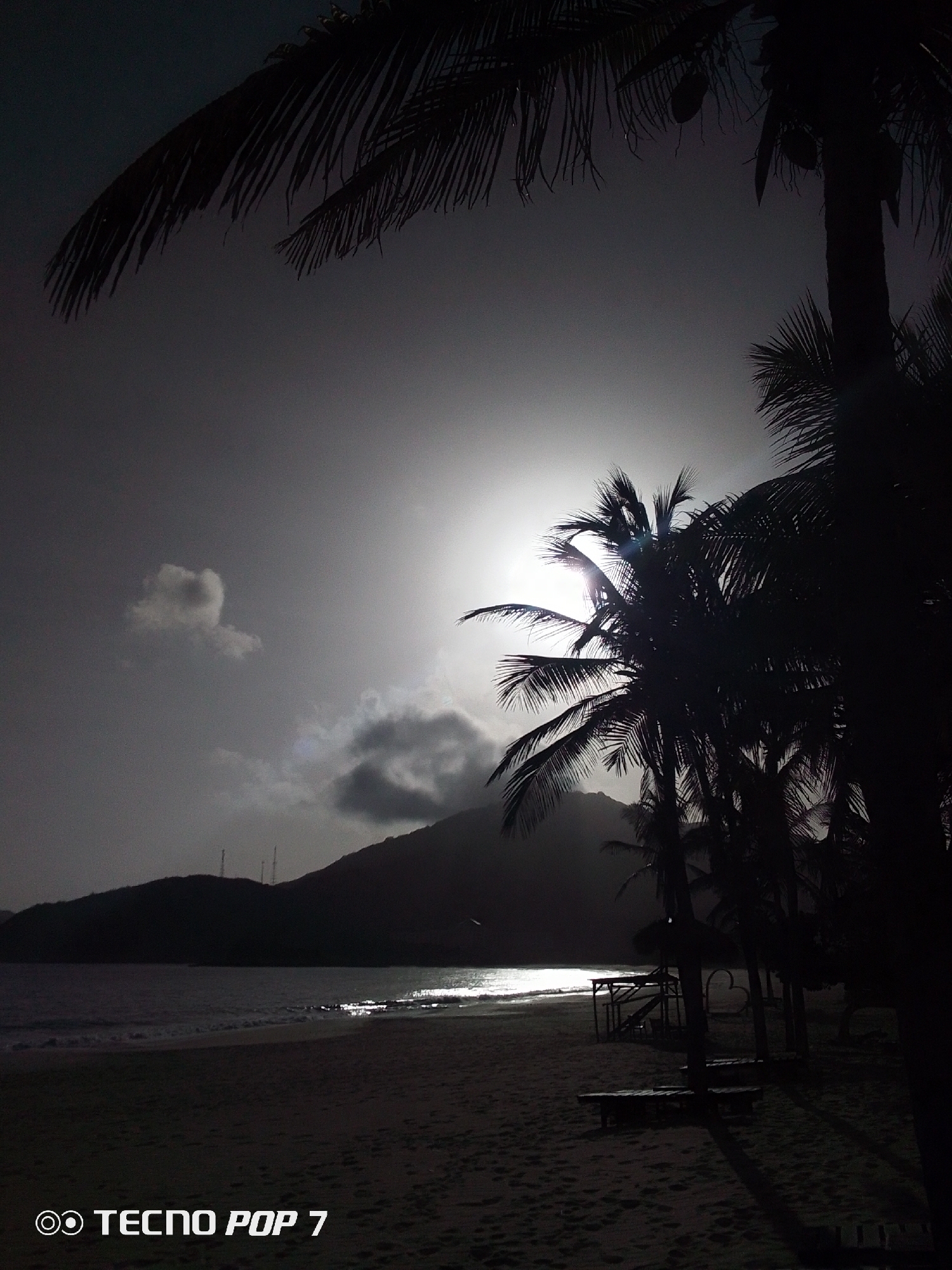 This is a low-light image of a beach in Pedro González, Venezuela. The beach appears deserted, with sand in the foreground and water stretching to the horizon. Dark silhouettes of palm trees frame the scene. The sun is partially obscured by clouds, creating a bright glare on the water. In the distance, the silhouette of a hill or mountain is visible. There are structures present in the middle ground, possibly seating areas or small buildings on the beach. The words "TECNO POP 7" are displayed at the bottom of the image.