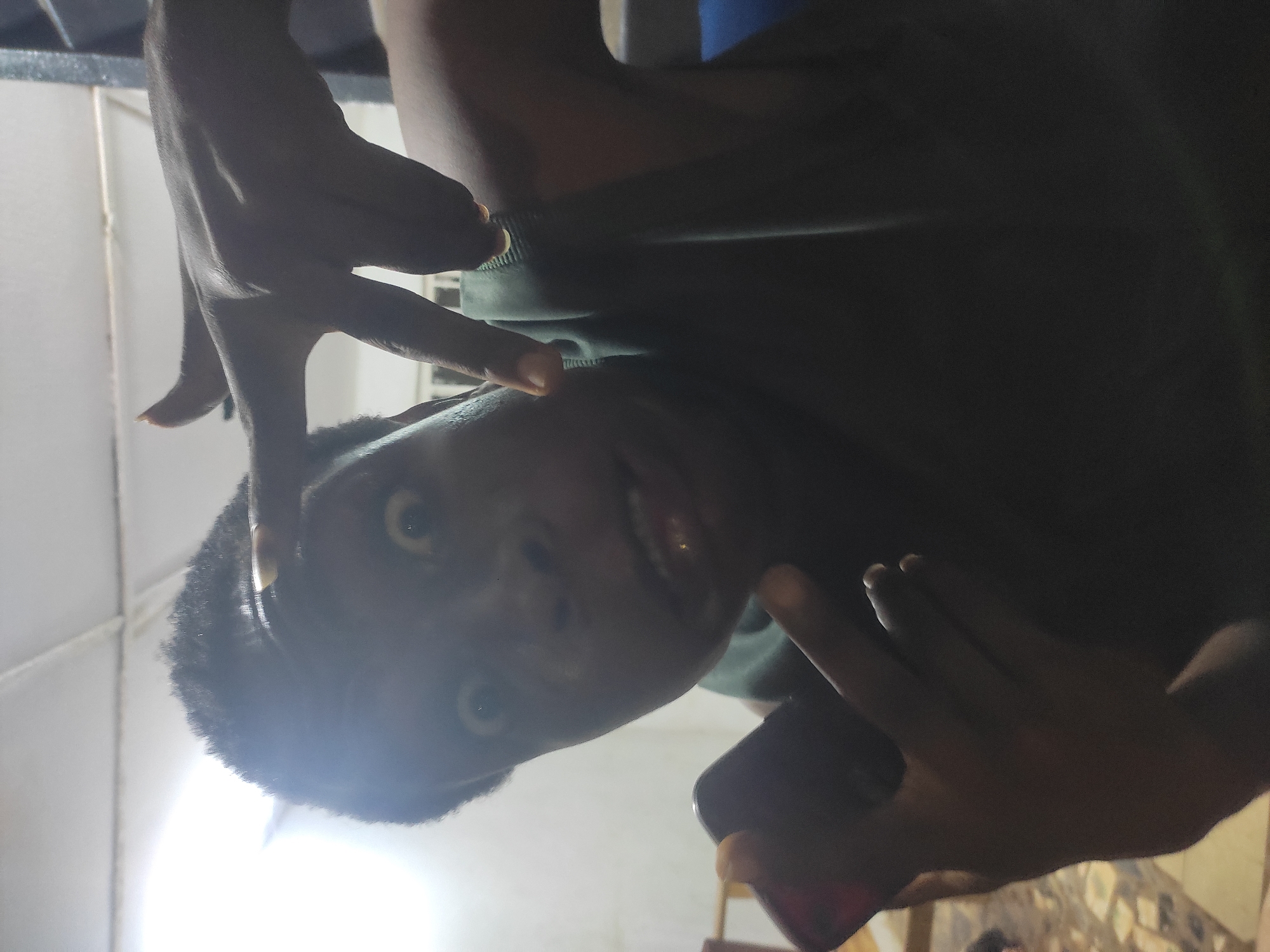 A medium shot depicts a dark-skinned person with short, black hair. The subject is holding their hands up, framing their face. The background shows tiled walls and flooring, suggesting an interior setting. The image appears to be taken from a low angle, tilted, and poorly lit. There is no definitive evidence in the image to confirm it was taken in Ikorodu, Nigeria.