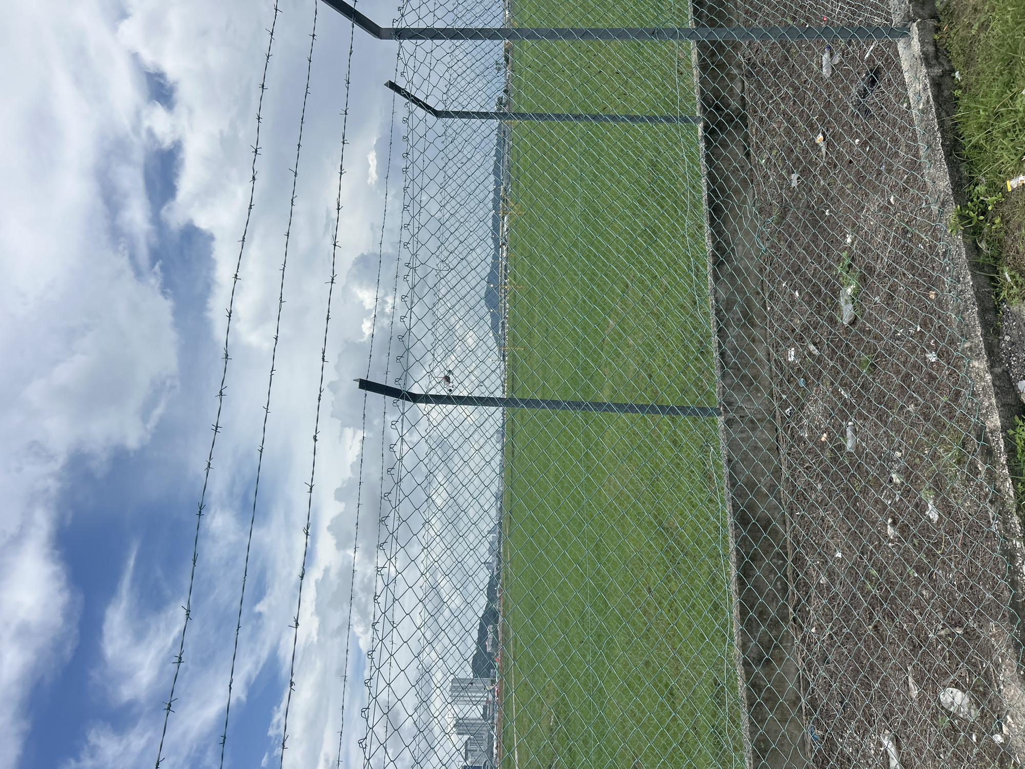 The image depicts a view of a fenced-off grassy field with barbed wire overhead. In the background, a city skyline with modern buildings is visible under a cloudy sky. The scene appears to be an undeveloped area within the city of George Town, Malaysia, possibly a construction site or an open space adjacent to urban development. The foreground shows a chain-link fence with a concrete base, and the ground behind it is covered with debris and dry leaves. The overall impression is one of an urban fringe, a transition zone between the built environment and open land.