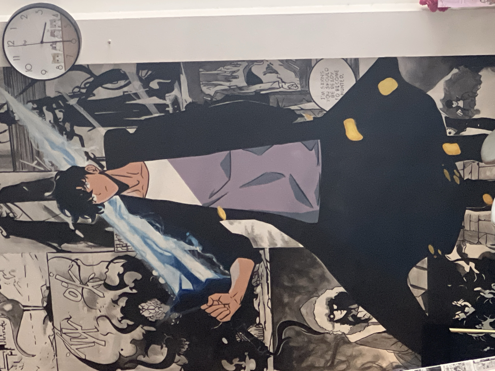 A low-angle shot captures a monochrome manga-style mural adorning a wall beneath a white ceiling. A round analog clock with a white face and black numerals and hands is positioned on the upper left of the frame. The mural showcases a central manga character, who is depicted facing left with an intense expression. The character is partially concealed by a solid black shape with yellow accents. A pale blue energy or force emanates from the figure's hands and around the character's body. A speech bubble with the text "I'M SAYINS YOU SHOULO BE READY TO BECOME HUNTED" is visible. The location is likely Baan Lamai, Thailand.