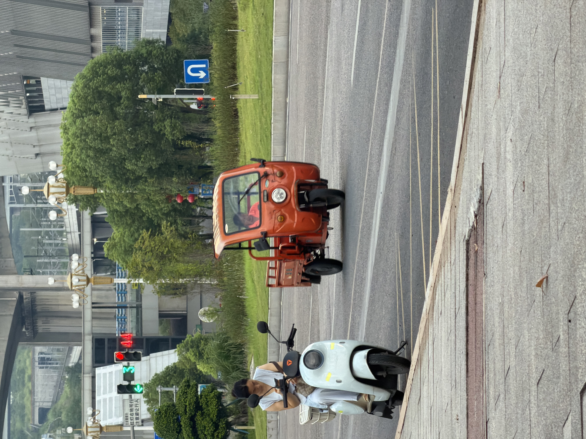 In the city of Jiangjin, China, a woman on a white electric scooter is looking at her phone while stationary at a traffic light. The scooter has a white basket on the back. To her right, an orange three-wheeled vehicle is moving away from the camera. The traffic light is red for the woman's direction, indicated by the red light illuminated above the green light. In the background, a large building with a modern facade is visible, along with lush green trees and overpasses. The ground is paved with concrete.