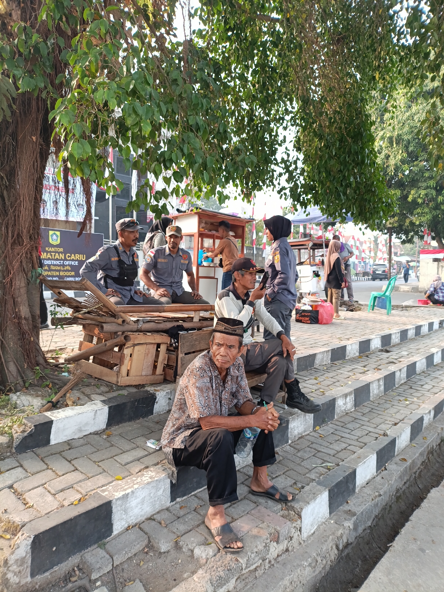 The image shows a street scene in the city of Cariu, Indonesia. In the foreground, two elderly men are sitting on a low wall beside a paved walkway. The man on the left is wearing a patterned shirt and black pants, and is holding a plastic bottle of water. The man on the right is wearing a black cap and is looking towards the left. Behind them, two men in grey uniforms with caps are sitting on a wooden structure. They appear to be members of a local official capacity, possibly law enforcement or public service workers. To their right, a food stall is set up with various items. In the background, there are more people walking, a car, and buildings. The scene is lit by natural sunlight, suggesting it is daytime. Red and white banners are visible, possibly indicating a local celebration or event. The presence of a large tree with hanging roots in the left of the frame adds to the natural environment within the urban setting. The ground is paved with interlocking bricks and bordered by curbs painted with black and white stripes.