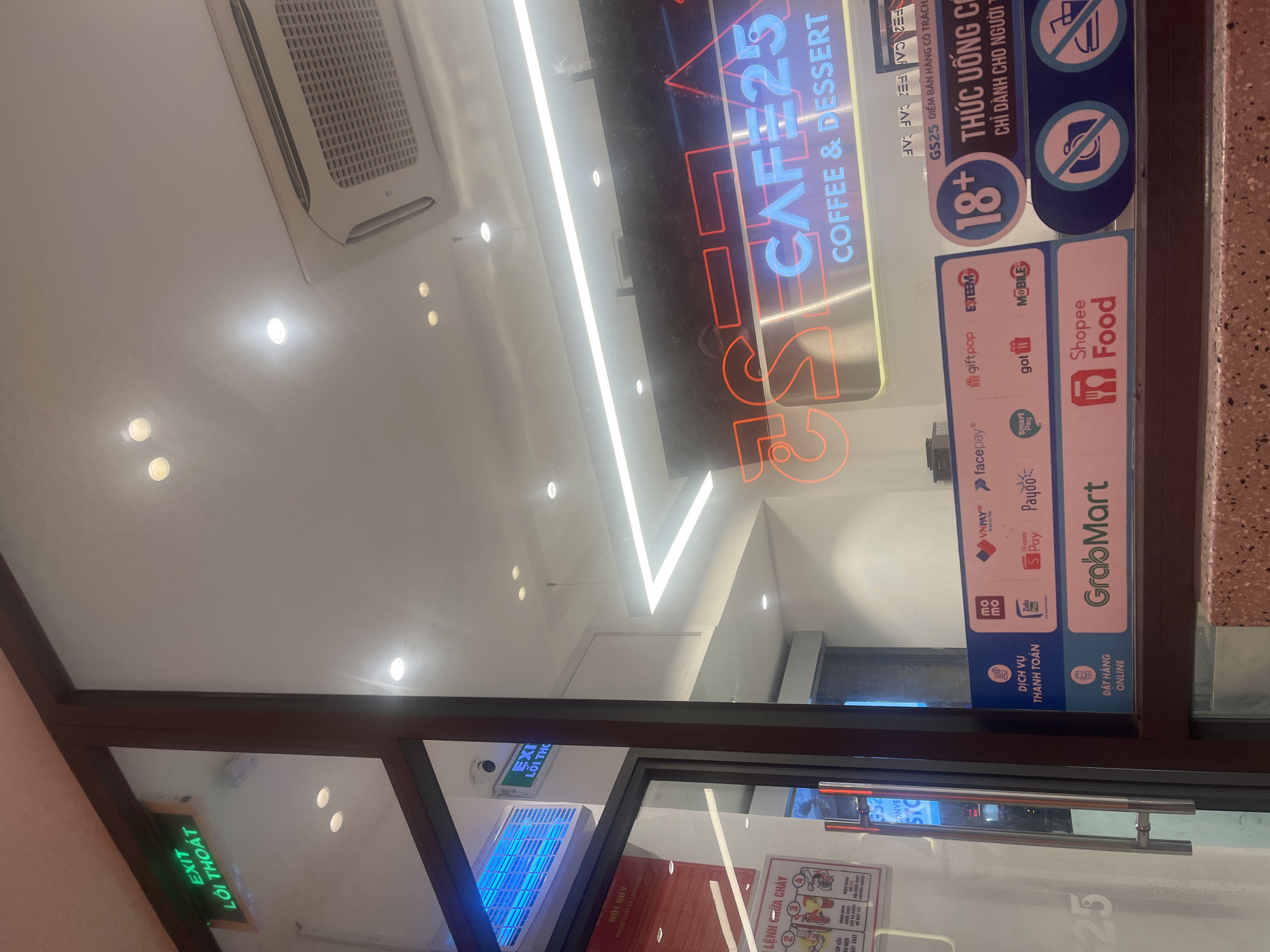 Here's a description of the image:
The image depicts the entrance of a Cafe25 location in Thủ Đức, Vietnam. The cafe offers coffee and desserts and has several delivery options, including GrabMart and Shopee Food. Payment options include Zalo Pay, MoMo, and Facepay. The cafe sells alcohol and beverages, and requires buyers to be 18+. The storefront has emergency exit signs with the Vietnamese translation. Regulations for preventing fire are posted.