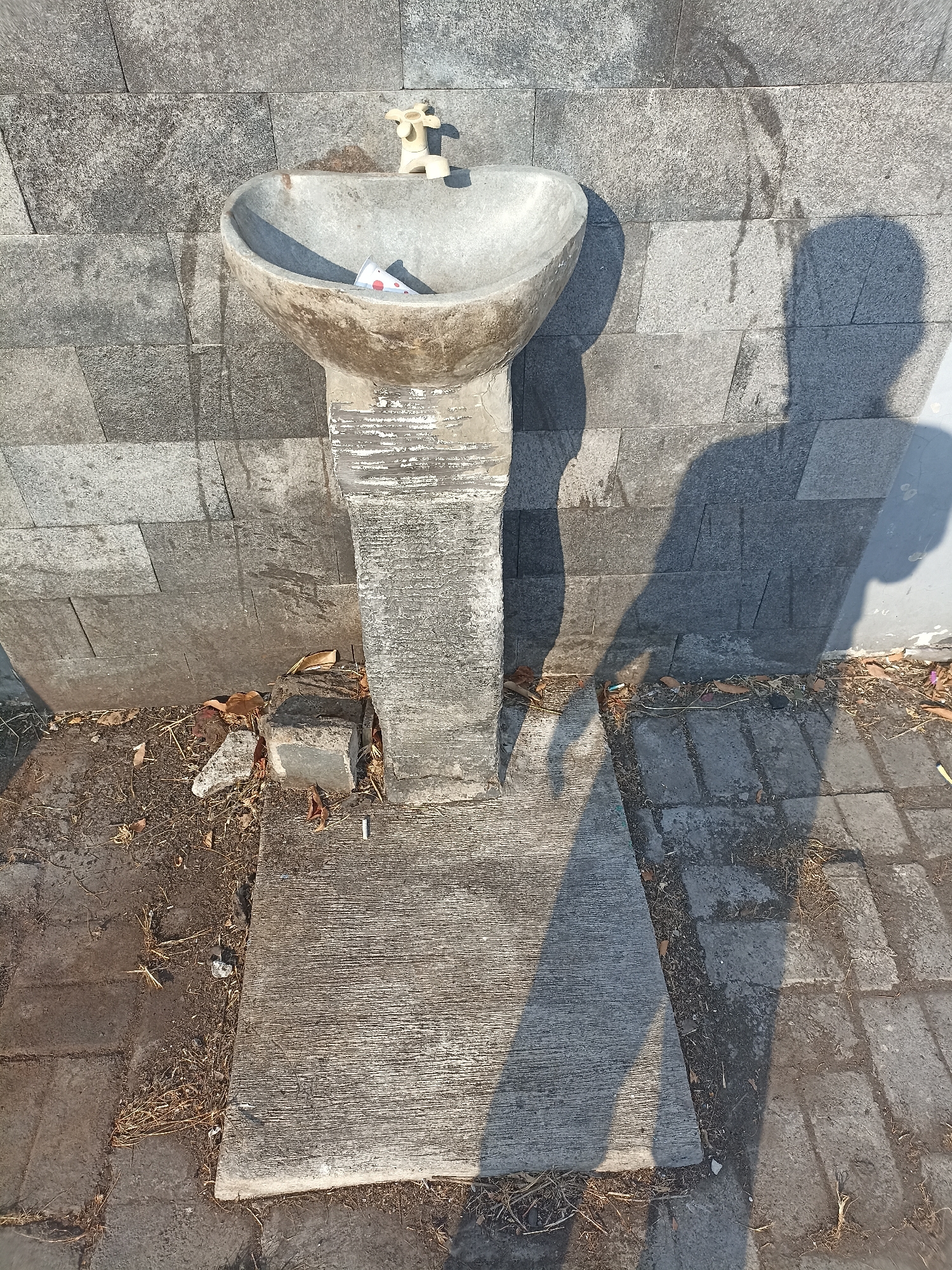 A close-up, high-angle shot captures a weathered, concrete washbasin with a light-colored faucet, situated against a gray, stone-tiled wall. A small, rolled-up piece of paper, possibly a cigarette pack insert, rests within the basin. The shadow of a person, cast by the sun from the right, engulfs a significant portion of the foreground and extends across the washbasin and wall. The ground beneath the washbasin is a mixture of concrete slabs and dirt, littered with dry leaves and debris, suggesting an outdoor, possibly public, area. A single cigarette butt is visible on the ground near the base of the washbasin. The scene appears to be an outdoor utility area, possibly in Mojopanggung, Indonesia, given the context provided. The overall impression is one of neglect and disuse.