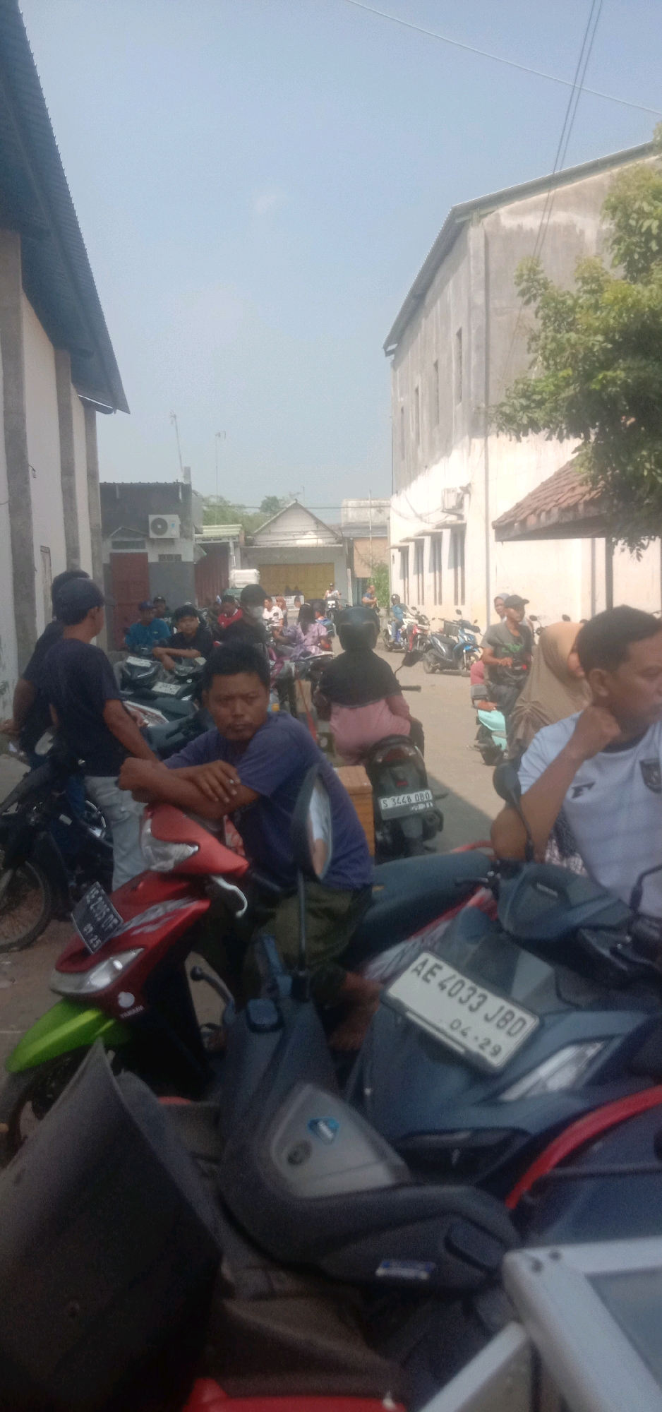 A group of people are gathered around motorcycles on a street in Cangkringrandu, Indonesia. The scene appears to be during the daytime, with sunlight casting shadows. Several motorcycles are parked, and some individuals are sitting on them, while others are standing and conversing. Buildings line both sides of the street, with one on the left appearing to be industrial or a warehouse, featuring a corrugated metal roof. The building on the right is a light-colored structure with multiple windows and a tiled roof. The sky is clear and bright blue. The overall impression is a common street scene with parked vehicles and people engaged in casual activity.
