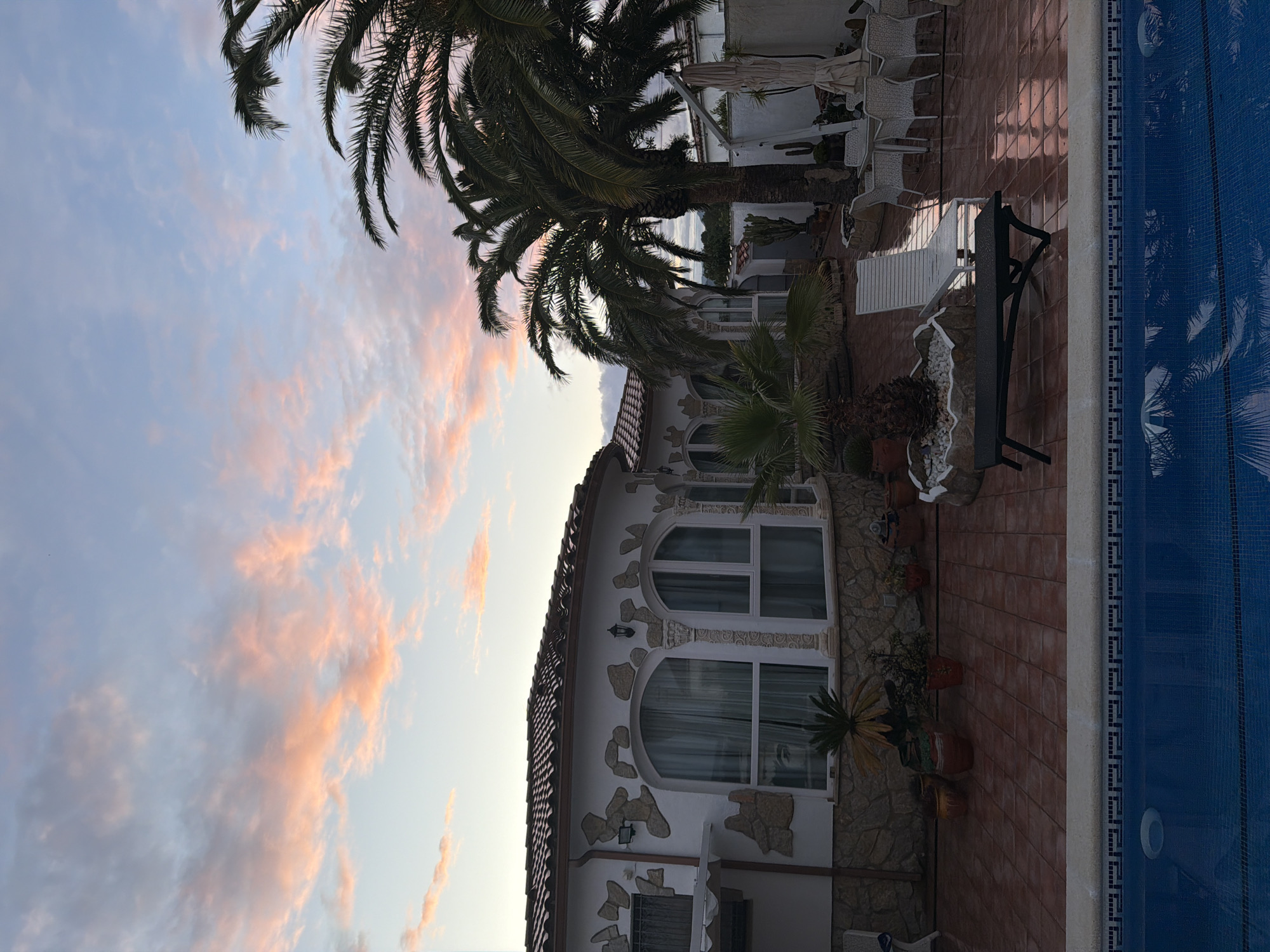 The image depicts a building facade in Mont-roig del Camp, Spain, next to a swimming pool. The sky is visible above the building, featuring a blend of light blue and pink clouds. Several palm trees are positioned to the left and above the structure, partly obscuring the view. The building itself has a white exterior with stone-like decorations around arched windows. A tiled patio leads up to the swimming pool, and a bench and various potted plants are placed along the patio. The swimming pool is lined with blue tiles and reflects the image of the trees.
