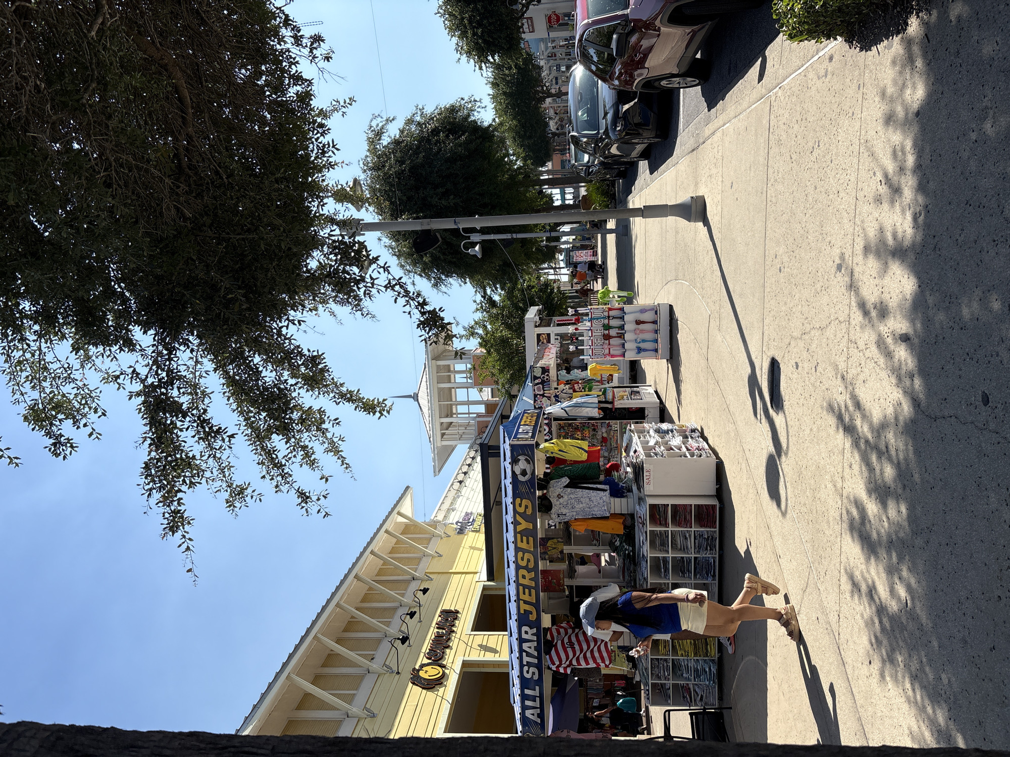 The image captures a sunny day in Panama City Beach, United States of America. The scene focuses on a sidewalk adjacent to a yellow building with the signage "Smile Just" and a smiley face. A merchandise stall labeled "ALL STAR JERSEYS" occupies the sidewalk, displaying sports jerseys and other items. A person, wearing a blue shirt, beige shorts, a cap, and sneakers, is walking along the sidewalk. Cars are parked on the street alongside the sidewalk, and trees provide some shade to the area. A light pole casts a shadow on the sidewalk. In the background, another merchandise stall and a raised, open-air structure are visible.