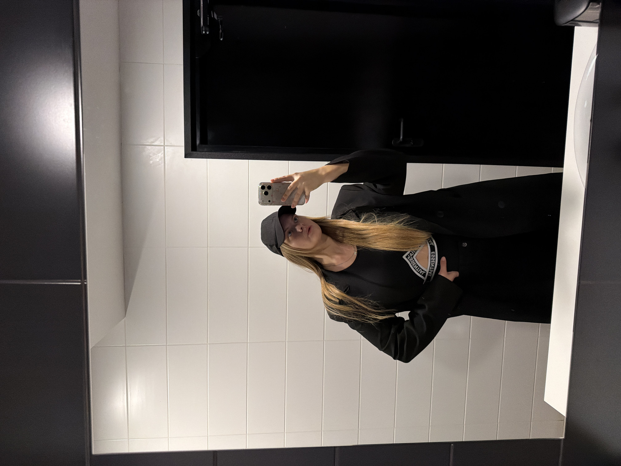 A woman is taking a selfie in a bathroom with white tiled walls and a dark countertop and fixtures. The woman is wearing a black baseball cap, a black shirt with white lettering around the neckline, and a black blazer. She is holding a cell phone in her left hand to take the photo. The bathroom has a dark-colored cabinet above the sink and is illuminated by bright light. The location is not identifiable as Izhevsk, Russia, or any other specific city based on the provided image.