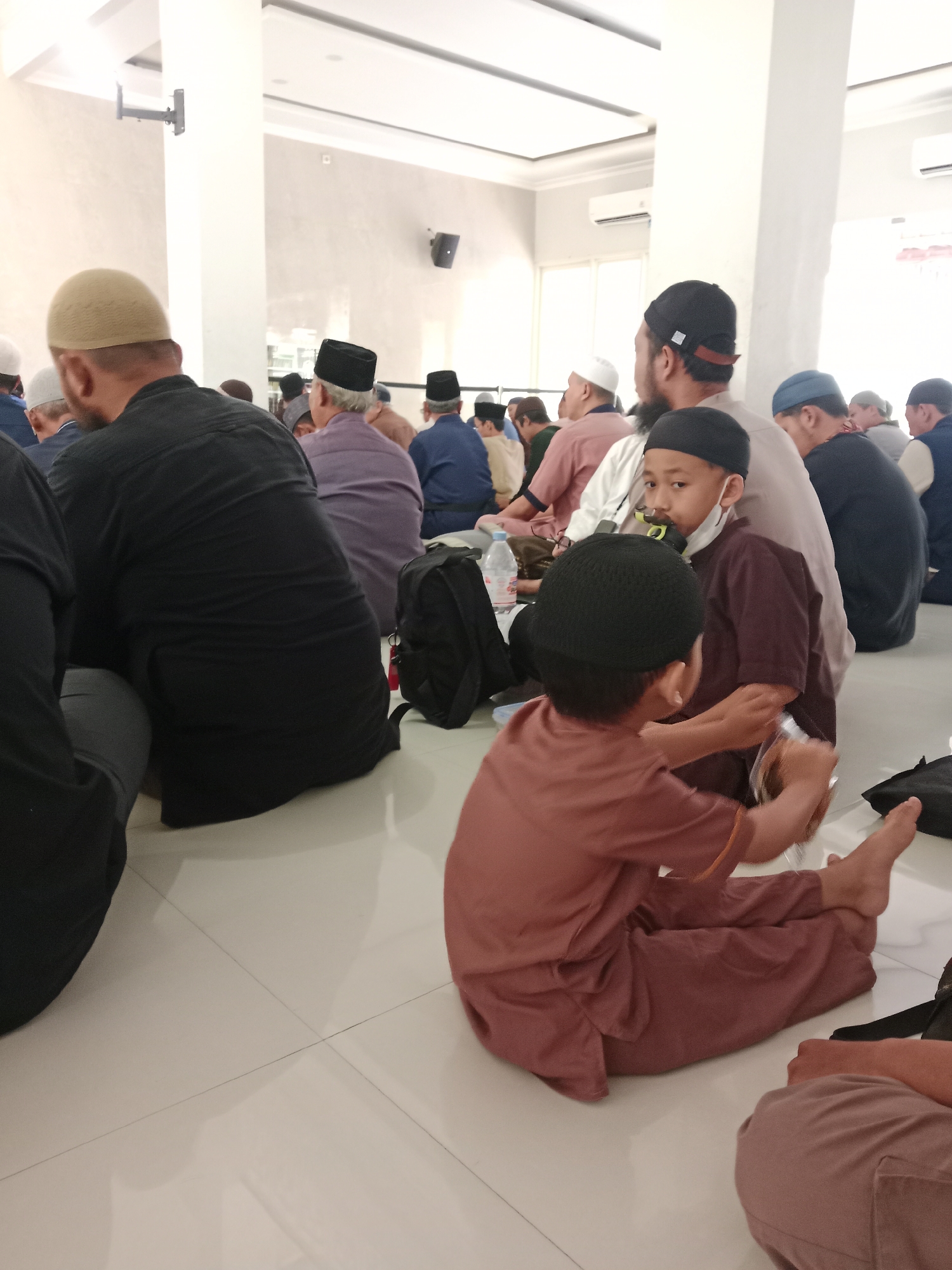 A group of men and boys, mostly wearing kufis, are sitting on a tiled floor inside a mosque, likely in Kediri City, Indonesia. The men are seated facing what appears to be the front of the mosque, while the boys are closer to the foreground. One boy in the foreground is wearing a brown shirt and pants and is looking away from the camera, with another boy in a similar outfit in front of him, also looking away. The environment is bright and appears to be well-lit with artificial lighting. There are air conditioning units visible on the wall. Some people are holding or interacting with small objects.