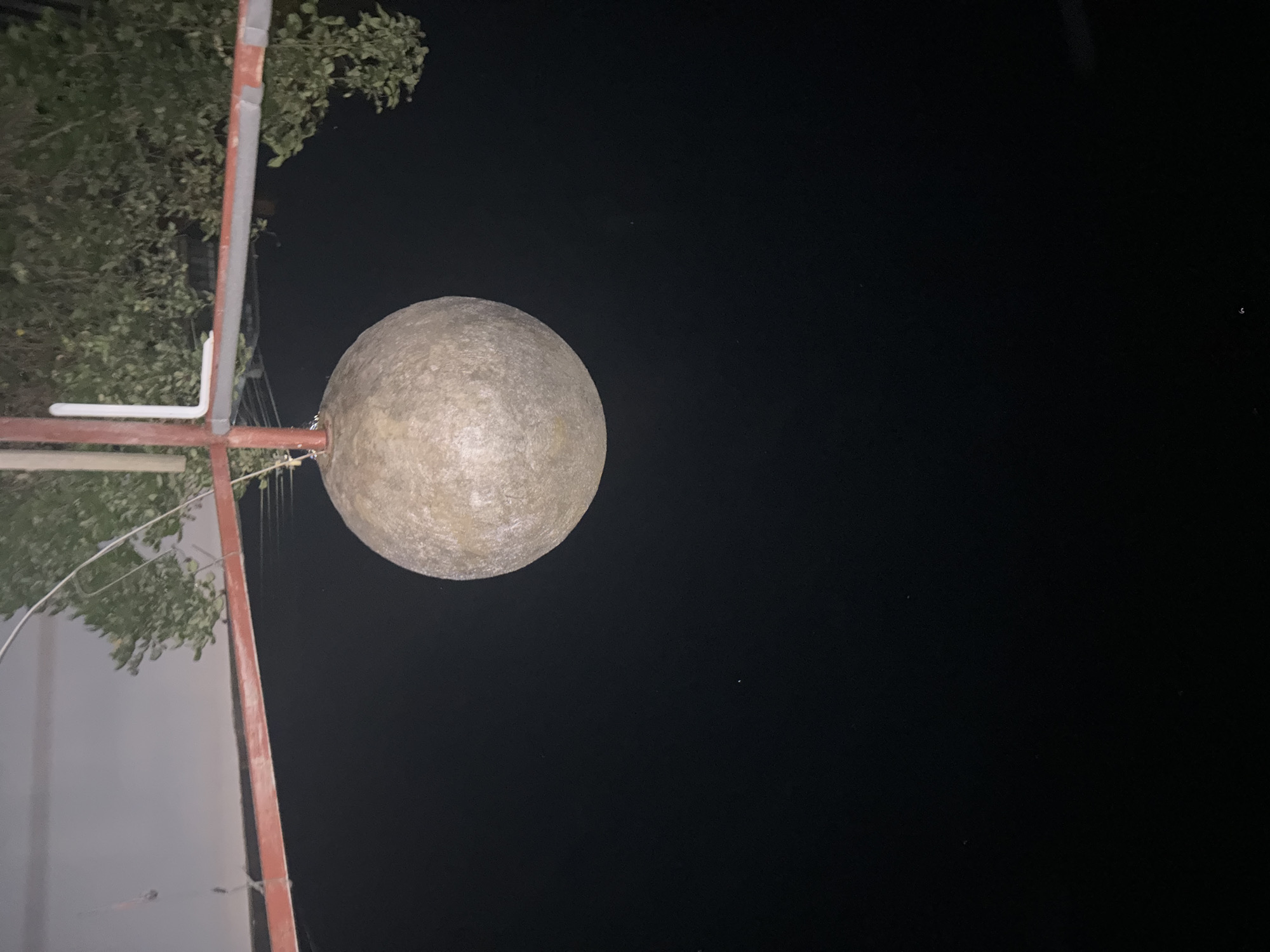 A low-angle, nighttime photograph shows a round, light-colored object mounted on a pole structure against a black background. The object appears to be some type of sphere or orb, possibly a lamp, attached to a red and grey metal pole. A few branches with leaves are visible in the upper left quadrant. The dark background suggests it is night. I cannot determine the location within Lom, Bulgaria.