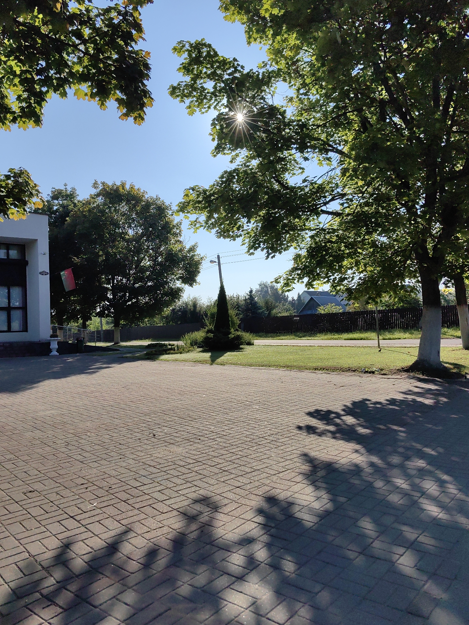 The image depicts a bright, sunny day in Fanipal, Belarus. In the foreground, a paved area made of rectangular, reddish-brown bricks is visible, with dappled shadows of tree leaves cast upon it. To the left, a white building with dark-paned windows and a decorative base is partially shown. A flag, red and green with some design, hangs from the building.
Further back, a lush green lawn stretches out, bordered by a dark wooden fence. Several trees with dense green foliage are scattered across the landscape. One prominent tree on the right side of the frame has a white-painted trunk, a common practice in some regions for tree health and visibility. A slender, cone-shaped evergreen tree stands near a lamppost. In the distance, houses with dark roofs and windows are nestled amongst more trees, suggesting a residential or semi-rural area within Fanipal. The sky is a clear, bright blue. The overall impression is one of a peaceful, well-maintained outdoor space.