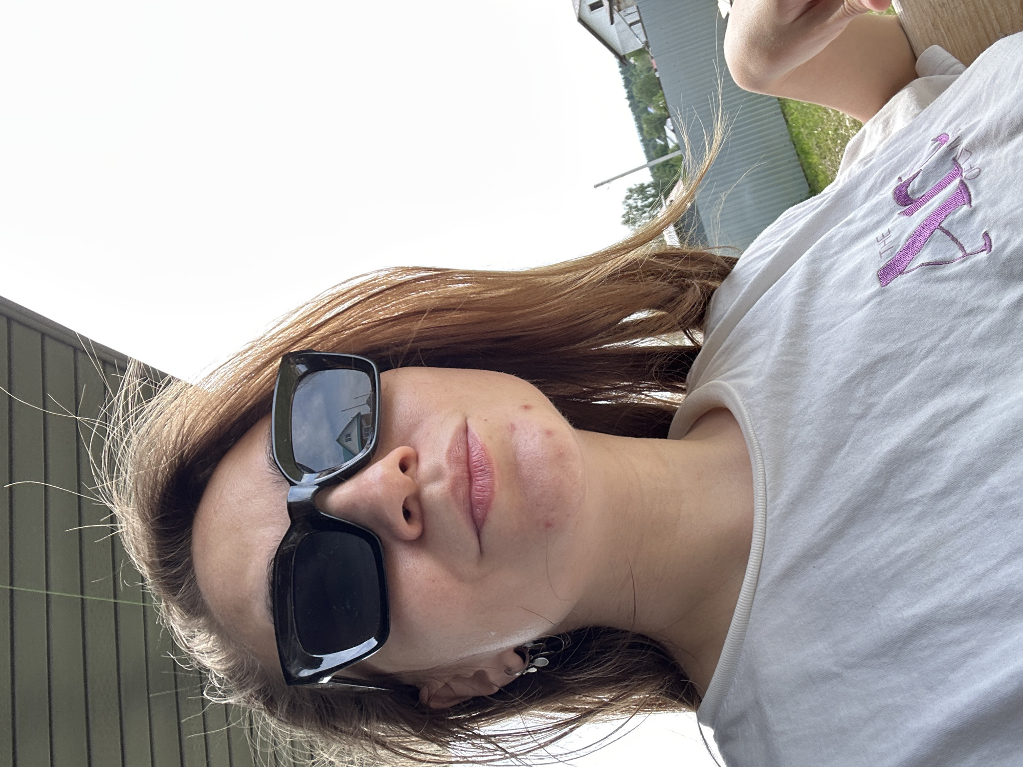 A woman with brown hair is wearing black sunglasses and a white t-shirt with purple embroidery. The picture is taken from a low angle, looking up at her face. Her head is tilted to the side. A building is partially visible in the background, and the sky is bright. There is no indication of the location being in Kharnavy, Russia.