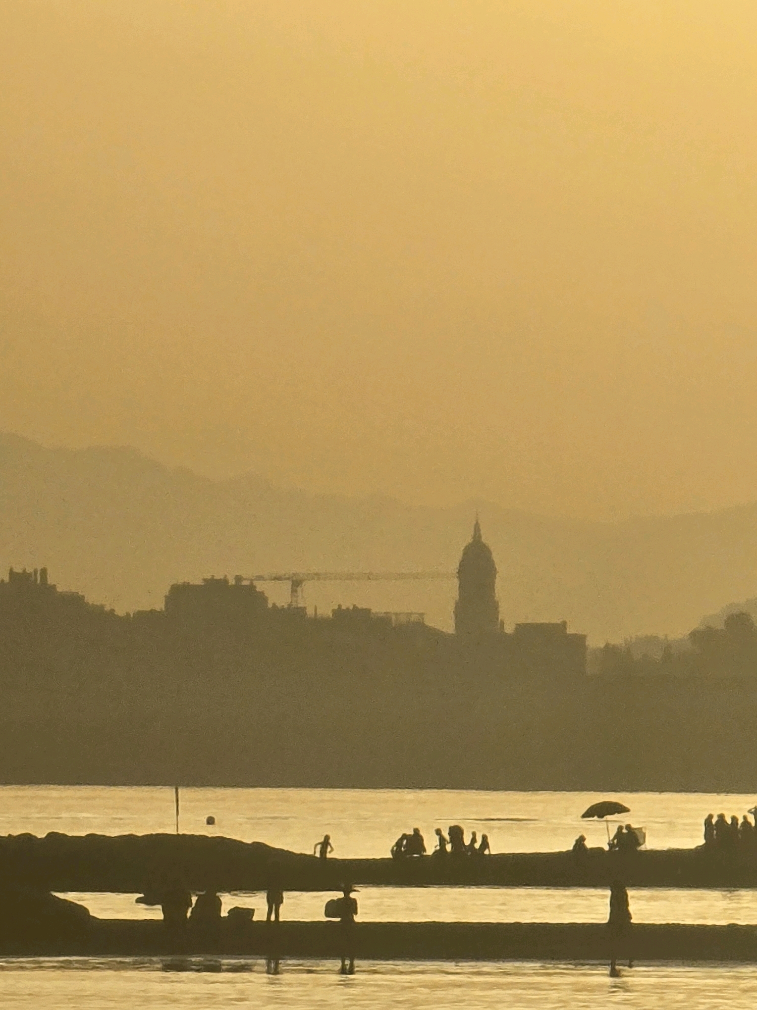 This is a silhouetted, low-angle, medium shot of a waterfront scene at sunset in Málaga, Spain. The golden hues of the setting sun dominate the image, casting a warm glow over the landscape and the water. In the foreground, a breakwater or jetty juts out into the calm water, with several silhouetted figures standing or walking on it. Some appear to be children playing, while others are adults gathered in small groups. One figure is walking through the water towards the viewer, and another is seated under a beach umbrella. The middle ground features the distinct silhouette of the Málaga skyline, with various buildings and a prominent clock tower. A construction crane is also visible within the city silhouette, suggesting ongoing development. In the background, hazy, layered mountains fade into the distance, further softened by the atmospheric conditions of sunset. The overall impression is one of a peaceful, late afternoon or early evening on the coast.