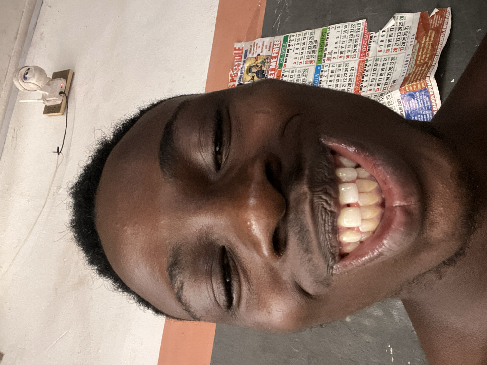 The image is a close-up selfie of a young Black male with dark skin, a wide grin revealing his teeth, and short, curly black hair. He is looking directly at the camera. In the background, a white security camera is mounted on a beige surface on a white wall. To the right of the subject, a portion of a calendar is visible, with the months of July, August, November, and December from the year 2026 printed on it. The calendar also displays dates, days of the week, and some text in a language that appears to be Igbo. The overall setting appears to be indoors. The presence of the calendar and the style of the calendar suggest the location is in Nkpor, Nigeria, a commercial hub known for its prevalence of such calendars and signage.