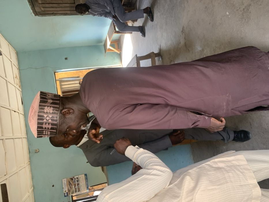 The image captures an indoor scene in Bojude, Nigeria, featuring four adult males. In the center-right foreground, a man with dark skin is seen from a three-quarter rear perspective, wearing a maroon long-sleeved traditional garment and a light pink cap with dark geometric patterns. His mouth is open, suggesting he is speaking. To his left, a man in a grey suit and white shirt is partially visible, facing slightly right with his left hand raised. In the immediate foreground left, the partial view of a person wearing a light-colored, striped long-sleeved garment is seen, with their right hand holding a pen near the left arm area of the man in the suit. In the background right, a fourth man is seated on a plain wooden bench, wearing a light grey long-sleeved traditional outfit and dark sandals, looking towards the left. The room features light blue painted walls, a white paneled ceiling with visible cracks and discoloration, and a concrete floor showing signs of wear. A poster or calendar is affixed to the wall behind the suited man. A dark wooden door is present in the background right, slightly ajar, revealing an illuminated area beyond. An empty wooden bench is also visible in the mid-ground. Natural light illuminates parts of the floor and the seated man in the background.