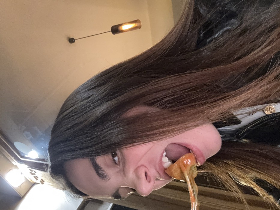 A young individual with long, dark brown hair is captured in a close-up, low-angle shot, actively consuming food within an indoor establishment in Paris, France. The subject's mouth is wide open, poised to bite into a piece of golden-brown, possibly fried or breaded, food coated in an orange-red sauce, presented on a fork. Their facial expression is intense, with eyes slightly narrowed and nose partially crinkled. The individual is wearing a black leather jacket over a lighter-colored garment, accessorized with a gold-toned circular pendant on a thin chain. The background reveals a light-colored ceiling with two artificial light sources: a bright, rectangular light panel or fixture on the upper left, and a single, warm-toned cylindrical pendant light hanging from the upper right. Vertical elements suggesting wall paneling or a window frame are also visible in the background.