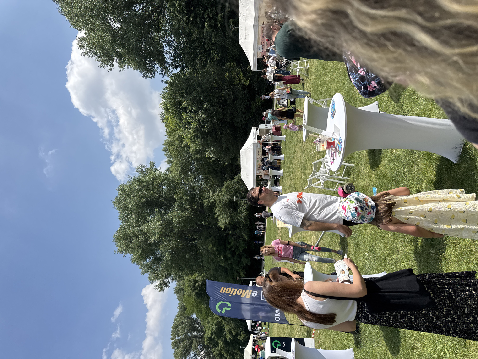 The image displays a bustling outdoor event or market taking place on a sunny day in a large park, identified as being in Sofia, Bulgaria, by linguistic cues on signage. The sky is bright blue with scattered white cumulus clouds. The ground is a lush, well-maintained green lawn, bordered in the mid-ground and background by dense, tall deciduous trees.
In the immediate foreground, a woman with long brown hair, wearing a white sleeveless top and a long black skirt with a white patterned design, is examining a pamphlet in her hands. A black fabric shoulder bag hangs from her left shoulder. To her right, a young child, facing away from the camera, wears a yellow dress with a lemon print and a colorful bucket hat adorned with dinosaur patterns.
Two tall, white, fabric-draped cocktail tables are positioned in the mid-foreground. One table holds a red cup, a clear cup with a straw, and several small informational flyers. To the right, partially visible, is the back of a person with long, wavy blonde or light brown hair, and further right, the back of a man wearing a dark green shirt.
Behind the foreground figures, a man with a beard, wearing a white t-shirt with red text and sunglasses on his head, looks towards the right. Behind him, a woman in a pink "Levi's" t-shirt and ripped blue jeans is visible, along with another man in a black t-shirt.
Multiple white canopy tents are set up across the grassy area, indicating vendor stalls or exhibition booths. On the left, a blue promotional flag features a green 'e' logo and 'eMotion' written vertically, with 'OMV' positioned below it. Adjacent to this flag, a charging station-like unit with OMV eMotion branding is visible. In the mid-ground to the right, one white tent clearly displays the Cyrillic text "PURISTO ПЕКАРНА" (Puristo Bakery), confirming the Bulgarian location. Numerous individuals of varying ages, including adults and children, are scattered throughout the park, walking, standing, and interacting, suggesting a lively community gathering. A baby stroller is visible near the rightmost tent.
