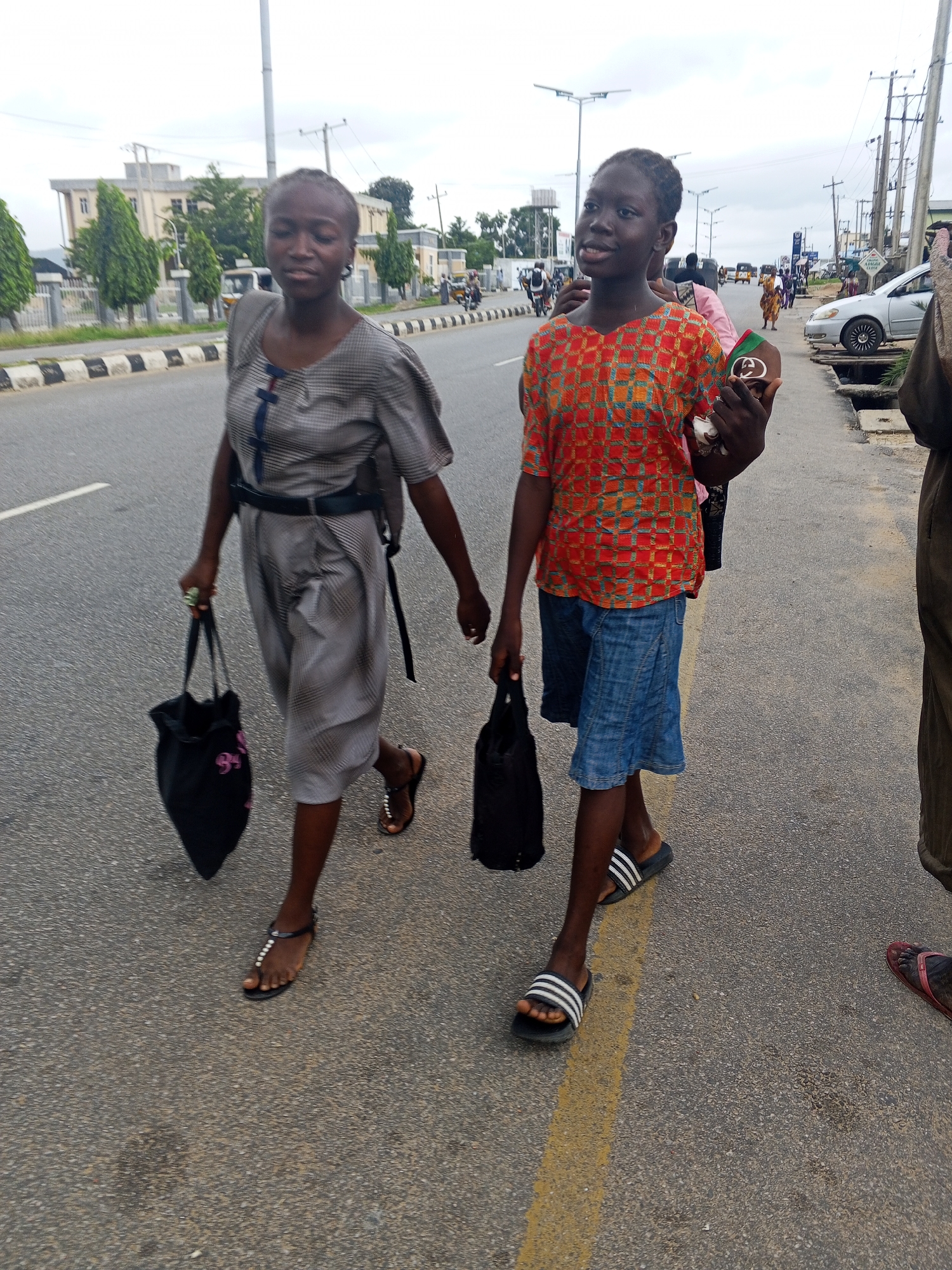 The image captures a street scene in Lokoja, Nigeria, likely during daylight hours given the ambient light. Two young women are the primary subjects, walking along the side of a paved road.
The woman on the left is wearing a grey, patterned dress with short sleeves and a dark belt cinched at her waist. She carries a black tote bag in her left hand and wears dark sandals adorned with white beads. The woman on the right is dressed in a colorful, patterned orange top and a blue denim skirt. She is holding a black tote bag in her left hand and wears black and white striped slides.
The background reveals a typical urban environment with a paved road marked with white lines. On the left side of the road, there is a sidewalk with a low black and white striped barrier. Beyond the sidewalk, buildings and trees are visible, including what appears to be a multi-story institutional building. On the right side of the road, another sidewalk is present, and further back, streetlights, utility poles, and various vehicles, including cars and motorcycles, are present, suggesting ongoing traffic and urban activity. The sky is overcast, indicating a cloudy day. The overall scene depicts pedestrian movement on a public street in Nigeria.