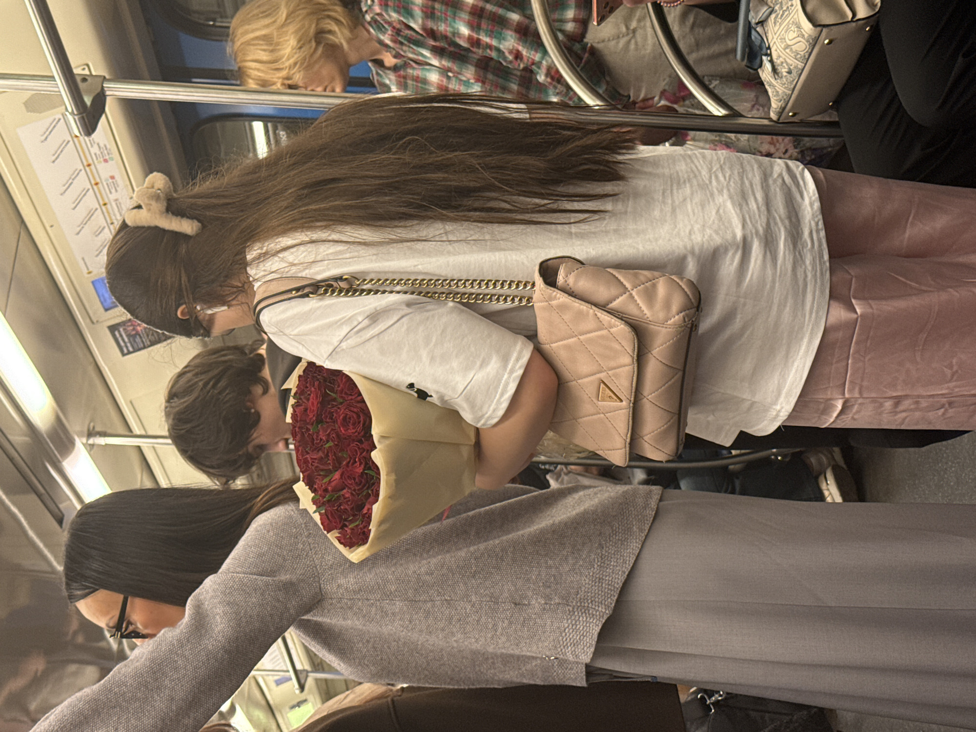 The image captures a scene inside a Moscow, Russia subway car. Several individuals are visible, including a woman with long brown hair and a clip in her hair, wearing a white shirt, pink pants, and a light pink purse. She is holding a bouquet of red roses, and is interacting with another individual. Other passengers include a person in a plaid shirt, and a woman wearing glasses and a grey outfit. The environment is typical of a subway car, including subway route maps and metal handrails.