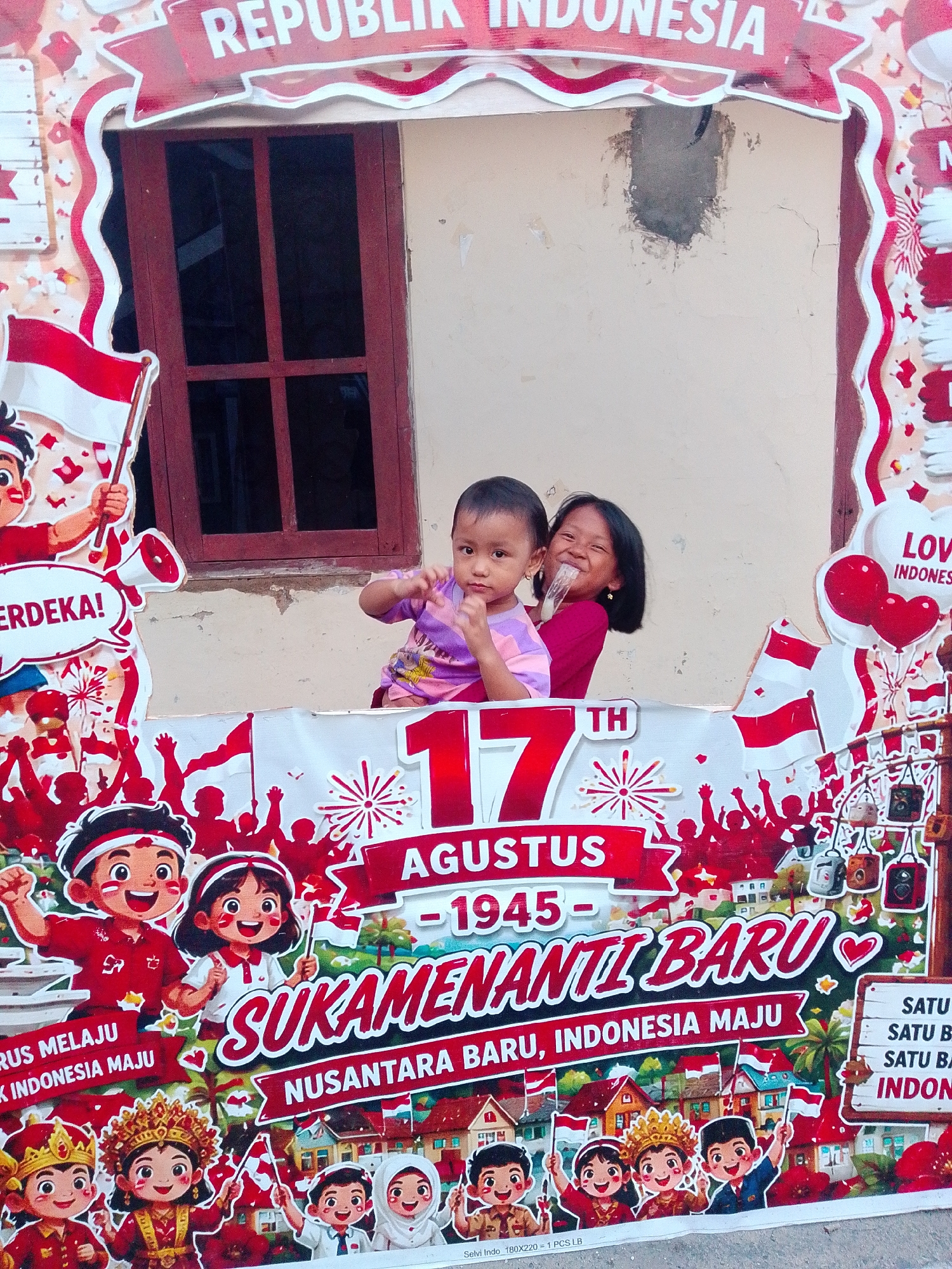 This is a medium shot photo of a photo booth with two young children posing behind a decorative cutout. The cutout is themed around Indonesian Independence Day. At the top of the cutout, "REPUBLIK INDONESIA" is written in white letters on a red banner. Below that, on the left, is a cartoon drawing of a child holding an Indonesian flag and a megaphone with the word "MERDEKA!" written in a speech bubble. On the right side of the cutout, a young boy is sitting, wearing a purple t-shirt, and a young girl is standing behind him, holding him and smiling widely, with a plastic cup in her hand.
Further down the cutout, the number "17" is prominent, followed by "AGUSTUS - 1945" and then "SUKAMANTIL BARU" written in large, stylized letters. Beneath that, it reads "NUSANTARA BARU, INDONESIA MAJU". The entire cutout is decorated with images of Indonesian flags, fireworks, a stylized cityscape, and more cartoon children in traditional Indonesian attire.
The background is a light-colored wall with a dark window. There are some visible cracks and patches on the wall. The photo booth itself is set up outdoors, likely in a public area, given the festive decorations. The lighting suggests it is daytime. The overall scene captures a moment of celebration and national pride, possibly related to an event or festival in Bandar Lampung, Indonesia.
