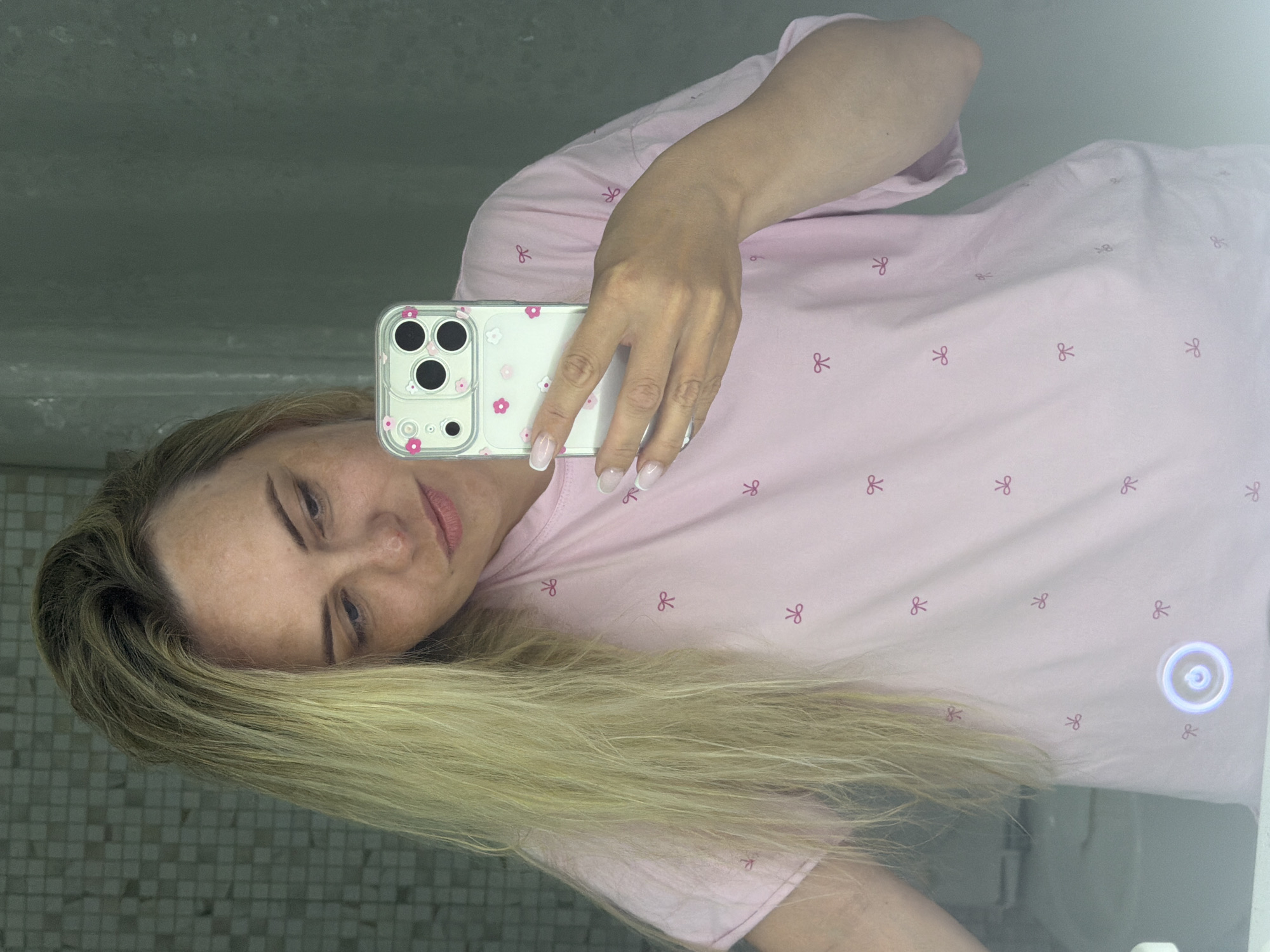 An image, likely a mirror selfie, shows a woman in what appears to be a bathroom setting in Votkinsk, Russia. The photo is rotated 90 degrees clockwise, so the woman's head is on the left and her body extends to the right.
The central subject is a woman with long, blonde hair, styled to fall over her shoulders. She has light makeup, including defined eyebrows and pinkish lips. She is wearing a light pink t-shirt adorned with small, repeating dark pink bow patterns. Her right arm is raised, holding a white iPhone with a clear case that features small pink and white floral designs. Her nails have a classic French manicure. She appears to be looking towards the camera within the phone, with a neutral expression.
The background, visible as a reflection in the mirror, shows a wall with small square tiles, possibly in shades of grey or green. To the right, on the mirror's surface, there is a circular, glowing button, typical of illuminated mirrors found in bathrooms.
The visible "text" from OCR seems to be misinterpretations of the bow patterns on the shirt (identified as "&" and "26") or noise. There is no clear, legible text in the image.