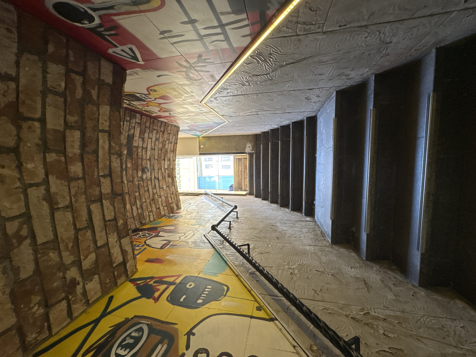 The image depicts an upward-sloping, interior stairwell or passage leading to an urban exit. The stairwell features dark, non-slip steps with lighter-colored anti-slip strips. The lower portions of the walls are composed of light grey, textured panels with a wavy or relief pattern. Above these panels, the walls are covered in vibrant graffiti artwork. The left wall displays yellow-backed graffiti with abstract shapes, a design containing the word "EYE," and a stylized robot-like face. The right wall features graffiti on a red and white background, including abstract elements, a partial arrow graphic, and the fragmented text "SURE COFFEE." The ceiling is an arched construction of brown and reddish-brown bricks. A dark, twisted metal railing is affixed to the left wall, while the right wall features a sleeker railing with an integrated LED strip on its underside, emitting a warm yellow light onto the textured wall below. The top of the stairwell opens to a brightly lit exterior, revealing the lower facade of a building with windows reflecting a light blue sky. The text "МОСКВА" (Moscow) is partially visible above the exit opening on the right side, identifying the location as Moscow, Russia. No individuals are present in the scene.