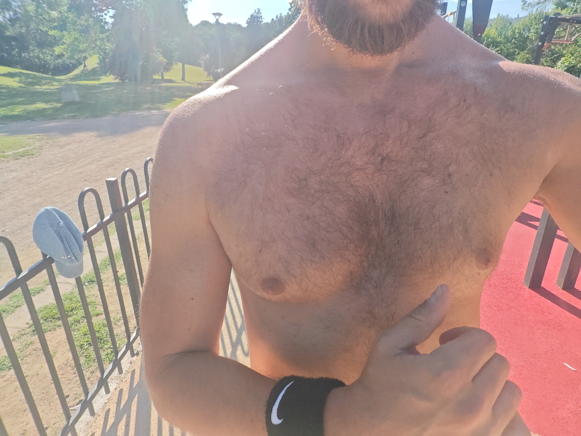 The media file shows a close-up of a shirtless person's chest and arm in an outdoor setting. The person has a beard and is wearing a black wristband with a white Nike logo. They are outdoors in what appears to be a park or recreational area, with a metal fence in the foreground and trees and a grassy hill in the background. The sun is shining, casting shadows and creating lens flare, suggesting it is a sunny day. The ground appears to be a mixture of dirt and a red rubberized surface, possibly a sports court. A light blue cap with white details is hanging on the fence. The overall impression is of a casual, outdoor activity, possibly exercise, given the athletic wear and park setting. The time of day appears to be daytime, with bright sunlight. The location cues, such as the vegetation and the style of the fence, are consistent with a park in Sofia, Bulgaria. There are no discernible emotions visible, as the face is not prominently featured. The only visible text is the Nike logo on the wristband.