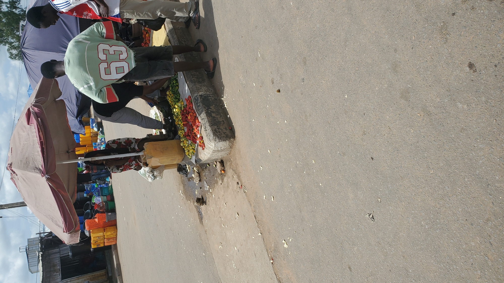 An outdoor, daytime scene depicts a street market in Jalingo, Nigeria, under a large, tan-colored umbrella providing shade. A woman wearing a floral-patterned top sits on a yellow jerrycan. In front of her, a display of produce, including red tomatoes and green peppers, is arranged on a raised concrete curb. Several individuals, appearing to be vendors or customers, are present. One man, wearing a light green sweatshirt with the number "63" on the back, stands to the right of the produce. Another man, in a white and red shirt, stands further to the right, facing away from the camera. The ground is paved with asphalt, with some debris and small puddles of water present. In the background, stalls with various goods, including stacks of yellow and orange plastic containers and buckets, are visible. The sky is partly cloudy.