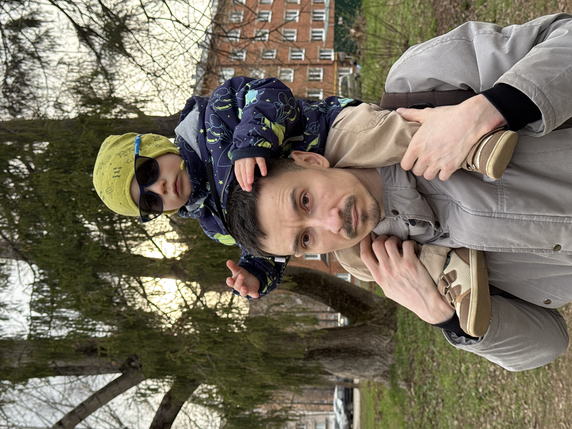 An outdoor, vertically oriented image captures a man carrying a young child on his shoulders, set against a backdrop of trees and a brick building, likely in Izhevsk, Russia.
The main subjects are an adult man and a young child. The man, with dark hair and a mustache, looks directly at the camera with a neutral expression. He is wearing a light grey jacket over a dark top and beige pants. His right hand is visible, holding the child's leg for support. The child is perched on the man's shoulders, wearing a bright yellow beanie with faint black text and drawings, and dark sunglasses. Their navy blue jacket features a playful pattern of light blue/green balloons or celestial bodies. The child's right hand rests on the man's head, and their left foot, clad in a beige and brown velcro shoe, is visible near the man's chest.
The activity depicts a parent-child interaction, possibly during a leisurely walk or while posing for a photograph. The child's sunglasses suggest bright daylight conditions, though the sky appears somewhat overcast or obscured by branches.
Notable details include the setting, which appears to be a park or residential area during late winter or early spring. Many trees have bare branches, while some conifers show green needles. A multi-story red brick building with numerous windows, characteristic of urban residential architecture, is visible in the middle ground. A dark-colored car is partially visible in the distant background. The ground shows patches of grass, some appearing dry. The text on the child's beanie is partially discernible, showing fragments like "dry" and "and."