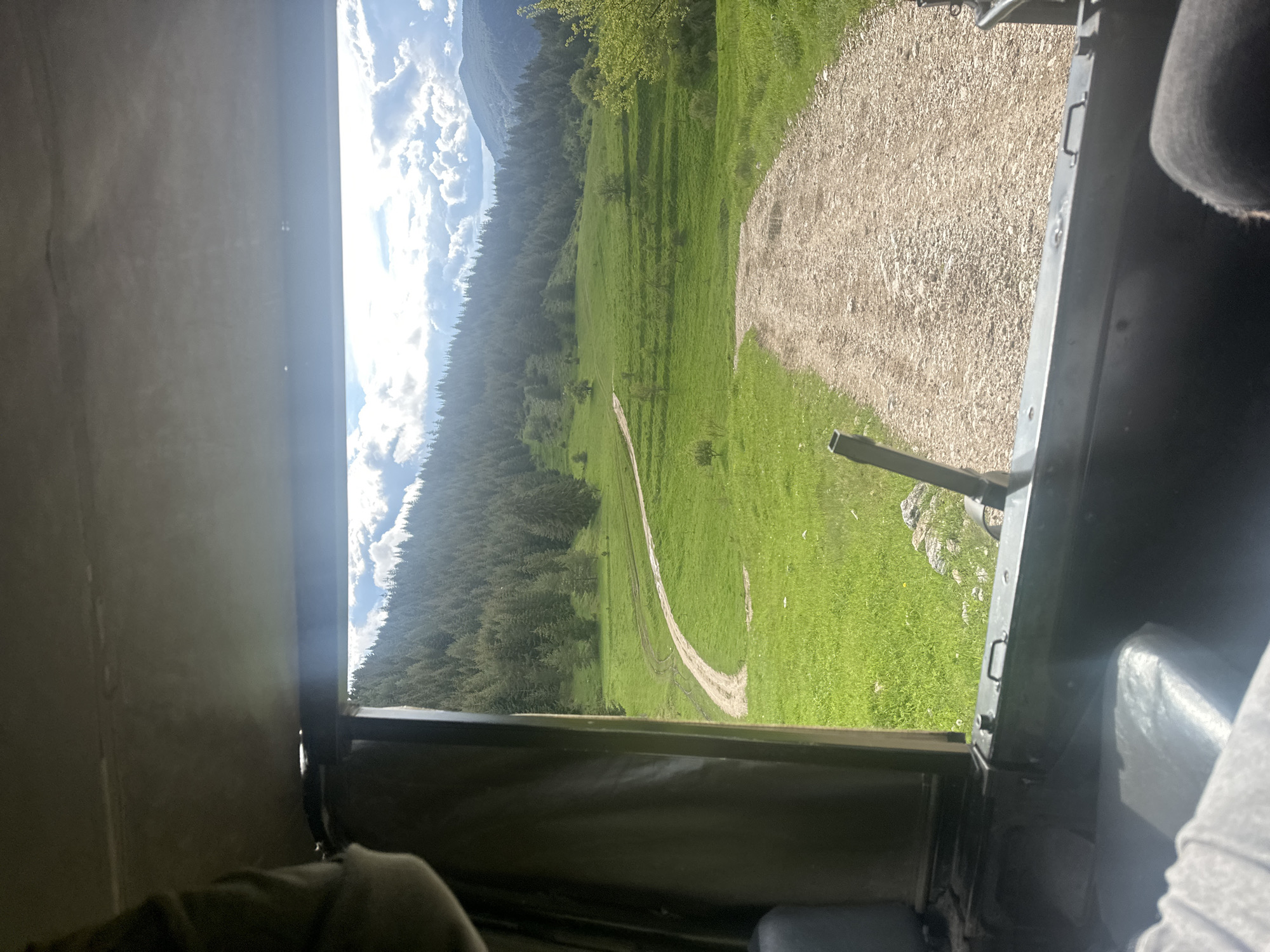 A view from inside a vehicle, likely a car or truck, looking out onto a rural landscape. A dirt road winds through a green meadow, with a dense forest on the left side of the frame. The sky is partly cloudy with mountains visible in the background. The interior of the vehicle, including parts of the door or frame, is visible in the foreground. There is no mention or indication of Iagodina, Bulgaria. Therefore, it is impossible to confirm its presence.
