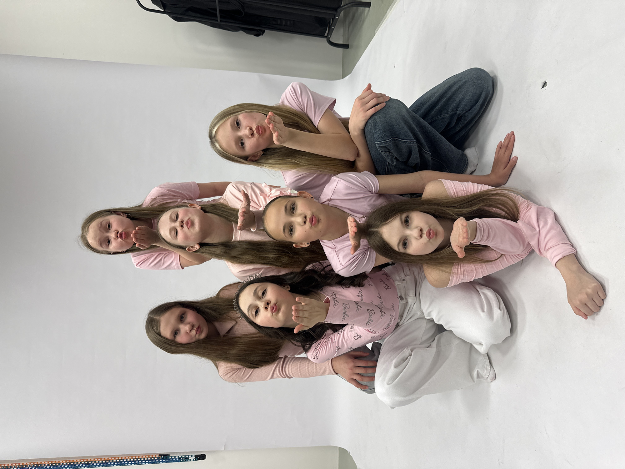 A group of seven young girls are arranged in a pyramid formation on a white surface. All the girls are wearing pink tops, and some have on jeans or white pants. All of them have their faces tilted upwards, and they are making kissing gestures with their lips and hands. The background is a simple white backdrop, possibly a photography studio. A black bag or equipment case sits in the top-left corner of the frame.
The image does not appear to be taken in the city of Votkinsk, Russia, but in an indoor photography studio.