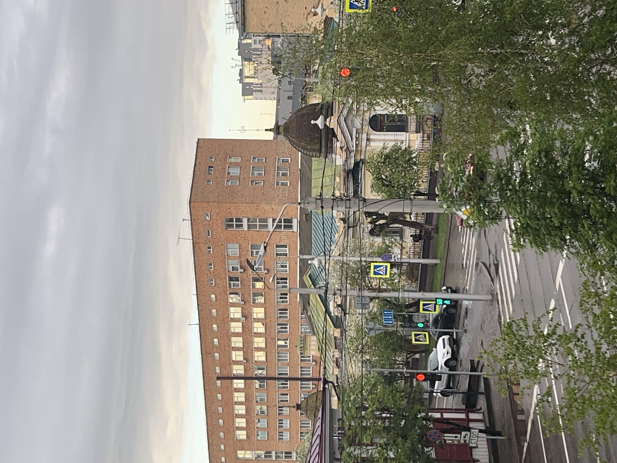 The image depicts an urban scene in Krasnoyarsk, Russia, viewed from an elevated position. A tall, reddish-brown brick building dominates the left side of the frame, with numerous windows. Adjacent to it, a domed building with architectural details is visible. A street runs through the right side of the image, complete with pedestrian crosswalks, street signs, and traffic signals. Two cars are present on the road. Trees frame the right side of the image. The sky is overcast.