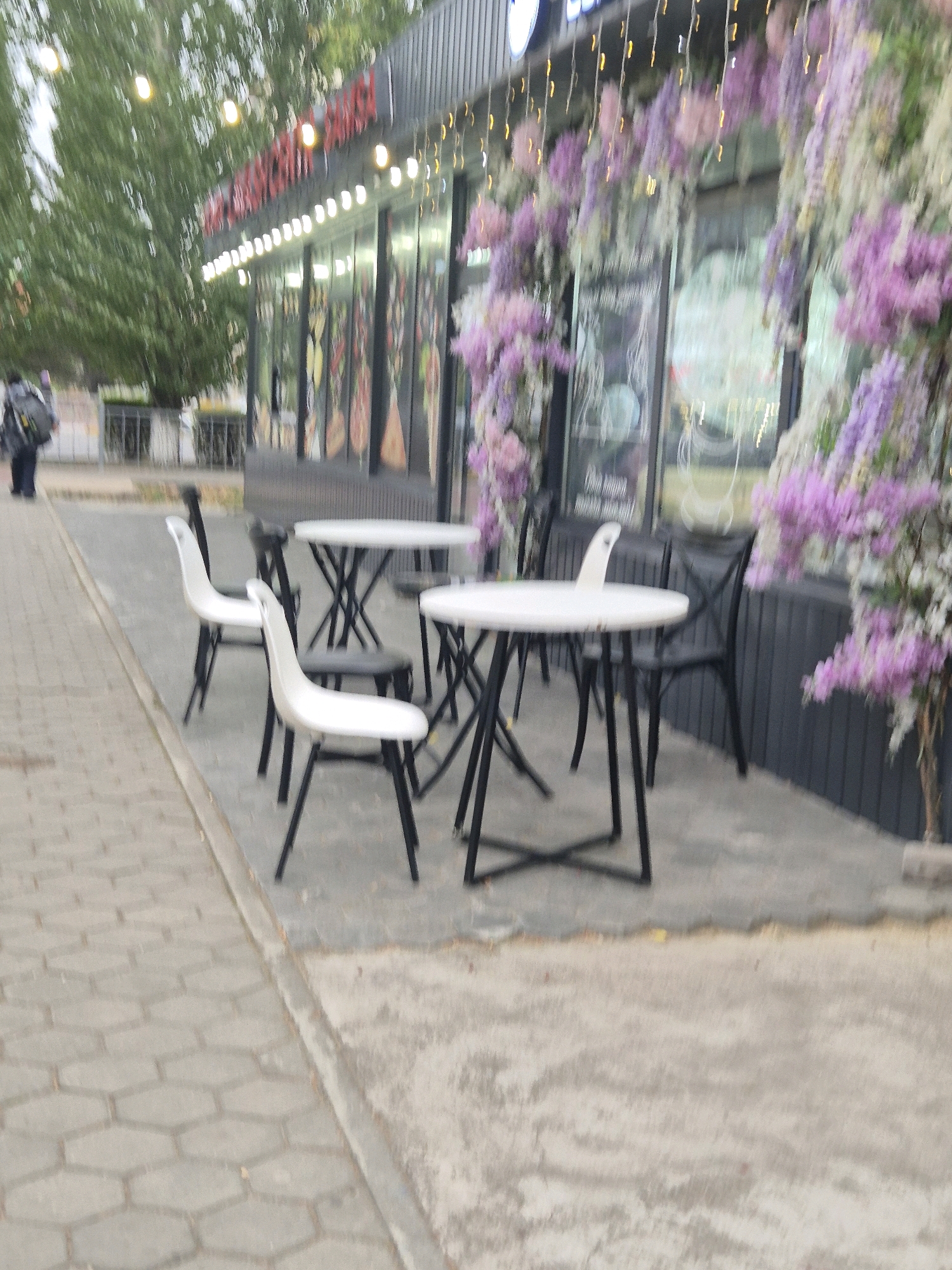 The image captures an outdoor seating area in front of a building, likely a cafe or restaurant, in Kokshetau, Kazakhstan. The foreground shows a gray, hexagonal paved sidewalk bordering a concrete patio. On the patio, several white tables with black metal bases are set up, accompanied by black chairs and some white chairs. The building facade features large windows and is adorned with an abundant floral decoration, predominantly in shades of purple and white, with string lights interspersed. A person carrying a backpack is visible walking on the sidewalk in the background to the left. The overall scene depicts a casual outdoor dining space in a city environment.