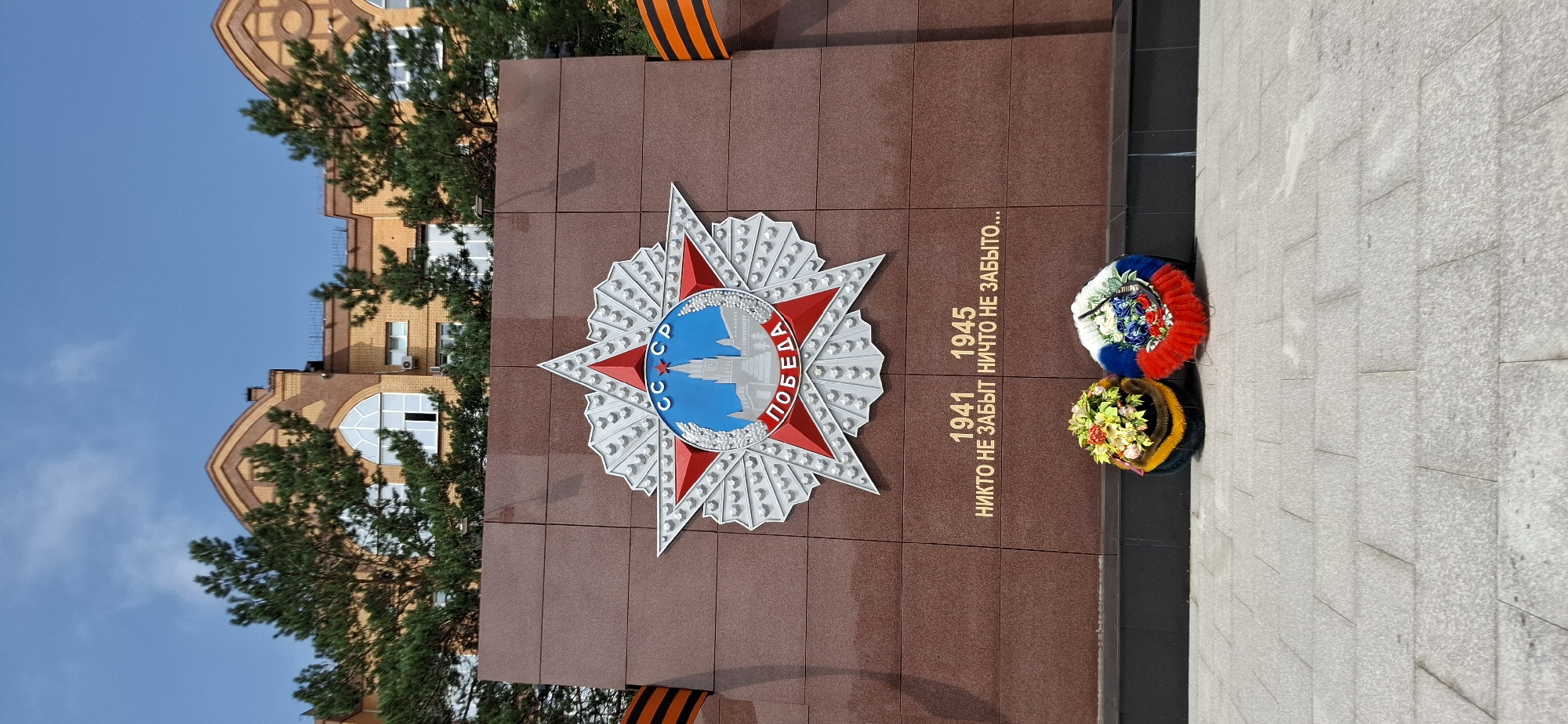 The image shows a monument with a large red star emblem at its center, featuring the letters "СССР" and "ПОБЕДА" in Russian. Below the emblem, the years "1941" and "1945" are inscribed, along with the phrase "НИКТО НЕ ЗАБЫТ НИЧТО НЕ ЗАБЫТО..." which translates to "No one is forgotten, nothing is forgotten...". Two wreaths are placed at the base of the monument; one is multi-colored with predominantly yellow and red hues, and the other features the colors of the Russian flag (white, blue, and red) with floral accents. Two orange and black ribbon decorations, resembling the St. George's ribbon, are visible on either side of the monument. In the background, a modern building with arched windows and brickwork is partially obscured by evergreen trees. The ground in front of the monument is paved with light-colored stone tiles. The overall scene appears to be an outdoor memorial site, likely in the city of Blagoveshchensk, Russia, commemorating a historical event, possibly related to World War II. No people are present in the image.