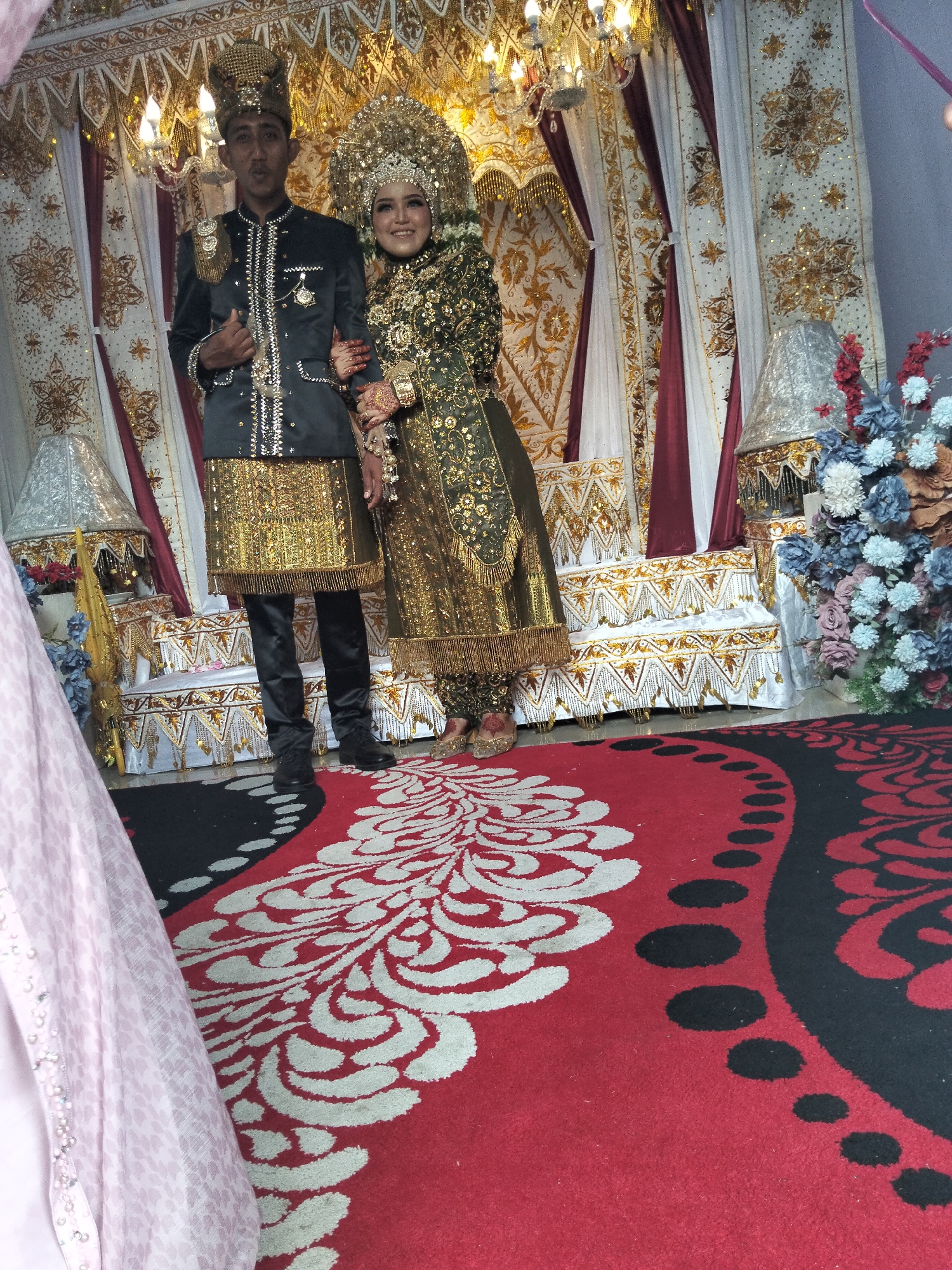 In a wedding ceremony taking place in Cottrueng, Indonesia, a newlywed couple stands adorned in traditional attire. The groom, on the left, wears a dark suit embellished with gold accents and a matching gold headpiece. The bride, on the right, is dressed in a striking green and gold traditional outfit, complete with an elaborate gold headdress. They are positioned in front of a richly decorated backdrop featuring gold and white drapes, and a chandelier hangs above. The floor is covered with a red carpet featuring a large white floral pattern. To the left, a pink fabric, possibly part of a dress or curtain, is visible. Decorative lamps and artificial flowers in shades of blue and red add to the festive ambiance.