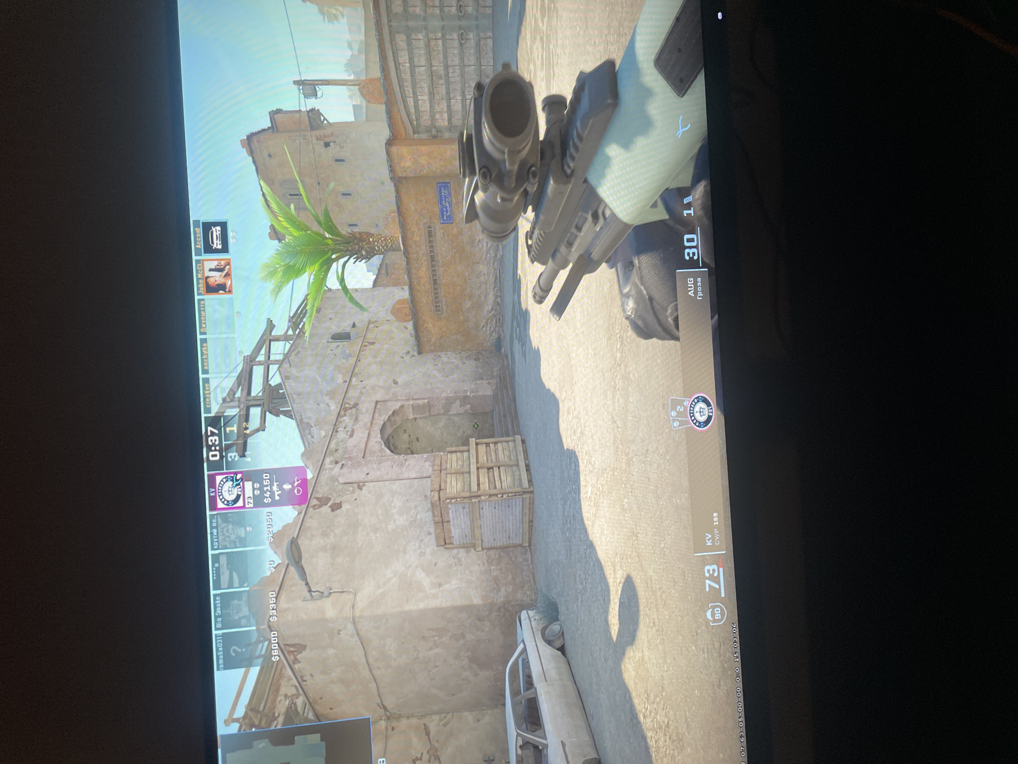 The image is a screenshot from a video game, likely Counter-Strike: Global Offensive, given the on-screen interface elements such as player names, scores, money, and timers. The scene appears to be a dusty, outdoor environment with weathered, beige buildings and a palm tree, suggesting a desert or Mediterranean-like setting. The perspective is first-person, showing the player's in-game weapon with a scope and ammunition counter.
On the left side of the screen, a player list is visible, showing player names like "romulia0311 Big Smoke," "anchуска Джульетта," and "John McCl...". Below these names are monetary values, indicating in-game currency. The timer shows "0:37" and the score is "3 1". On the right side, the player's rank is displayed as "90" and "73" with some text indicating "CV/P 159". There's also text in Russian, "AUG 30 1 Гроза," which translates to "August 30 1 Storm." The overall atmosphere is tense and action-oriented, typical of a competitive shooter game. The lighting suggests it is daytime. The presence of a car in the background and wooden crates hints at a combat scenario. The location context "Seversk, Russia" is provided externally and not visually evident in the screenshot itself.