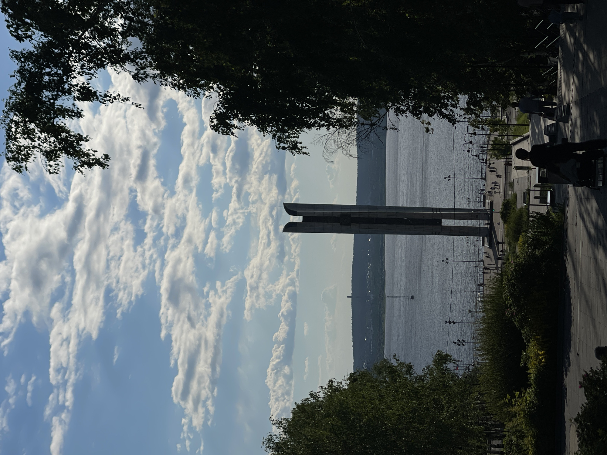 The image depicts a scenic waterfront promenade in an urban setting, likely during daylight with partly cloudy skies. The scene is framed by lush green trees on either side, casting shadows and creating a natural frame. In the center of the composition, a tall, modern monument stands prominently on the waterfront, characterized by its slender, segmented structure. The wide body of water, possibly a river or lake, stretches across the middle ground, with a distant, tree-lined horizon. Along the waterfront, a paved promenade is visible, populated by a few individuals walking and a boat in the distance on the water. A set of stairs leads down to the promenade from a higher vantage point, where several people, silhouetted against the bright sky, are descending. Streetlights line the promenade, suggesting it is a public area designed for leisure and viewing. The overall atmosphere is serene, highlighting the interplay between nature, urban architecture, and human activity.
