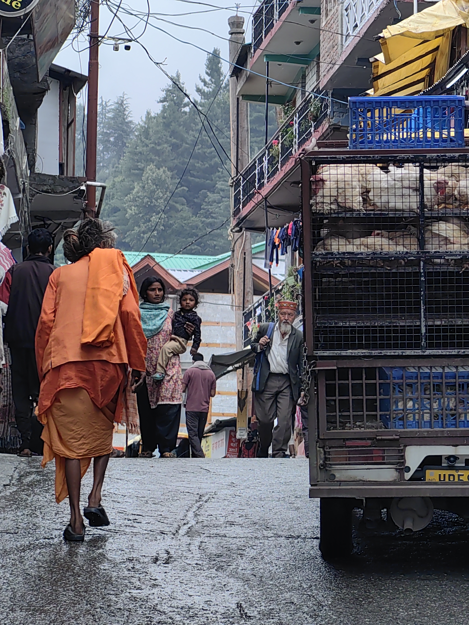 In Vashisht, India, on a wet, paved street, a man in saffron robes walks away from the camera, his back to the viewer. He wears dark sandals. To his right, a woman, holding a young child, walks towards him. The woman wears a floral patterned top and a blue scarf. The child is dressed in a dark top and beige pants. Further to the right, an elderly man with a white beard and wearing a dark jacket and a patterned cap walks down the street. He carries a blue backpack. In the foreground right, a portion of a truck is visible, loaded with cages containing white fowl. The street is lined with buildings, some with balconies, and overhead wires are prevalent. Trees are visible in the background. The overall atmosphere appears to be overcast.