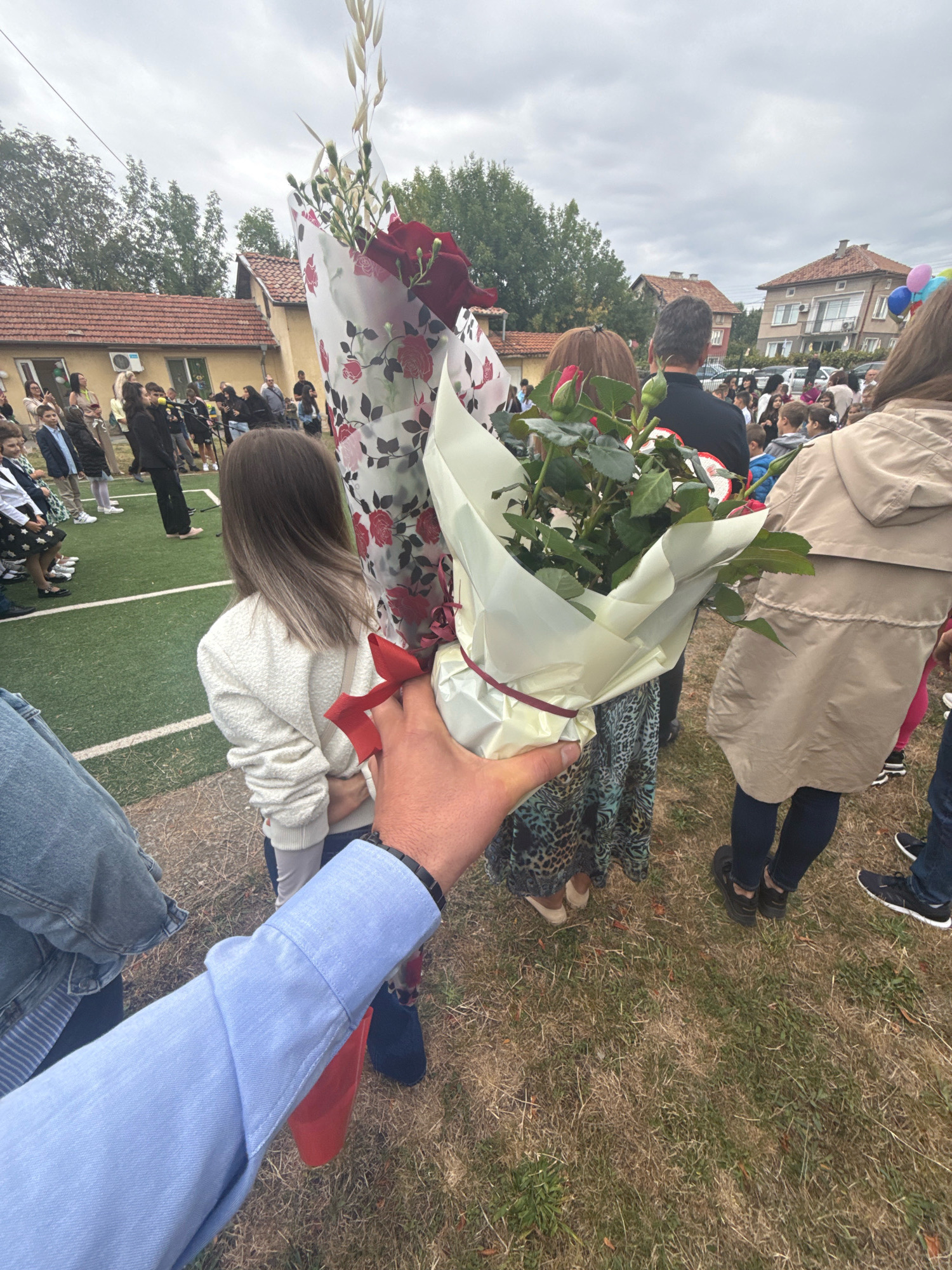In Kostinbrod, Bulgaria, on what appears to be the grounds of a school, a person's hand, clad in a light blue dress shirt, holds a bouquet of roses and dried wheat stalks wrapped in decorative paper. The bouquet is positioned in the foreground, obscuring part of the scene. Behind the bouquet, a young girl with shoulder-length brown hair, wearing a cream-colored textured sweater, is seen from the back. She is standing on a grassy area adjacent to a green artificial turf field. Several other individuals, mostly children and adults, are gathered in the background, some standing on the field and others on the grass. Buildings with tiled roofs are visible beyond the gathering, suggesting an urban or suburban setting. The sky is overcast. The overall scene depicts an outdoor event, possibly a school ceremony or gathering, given the presence of children and flowers.
