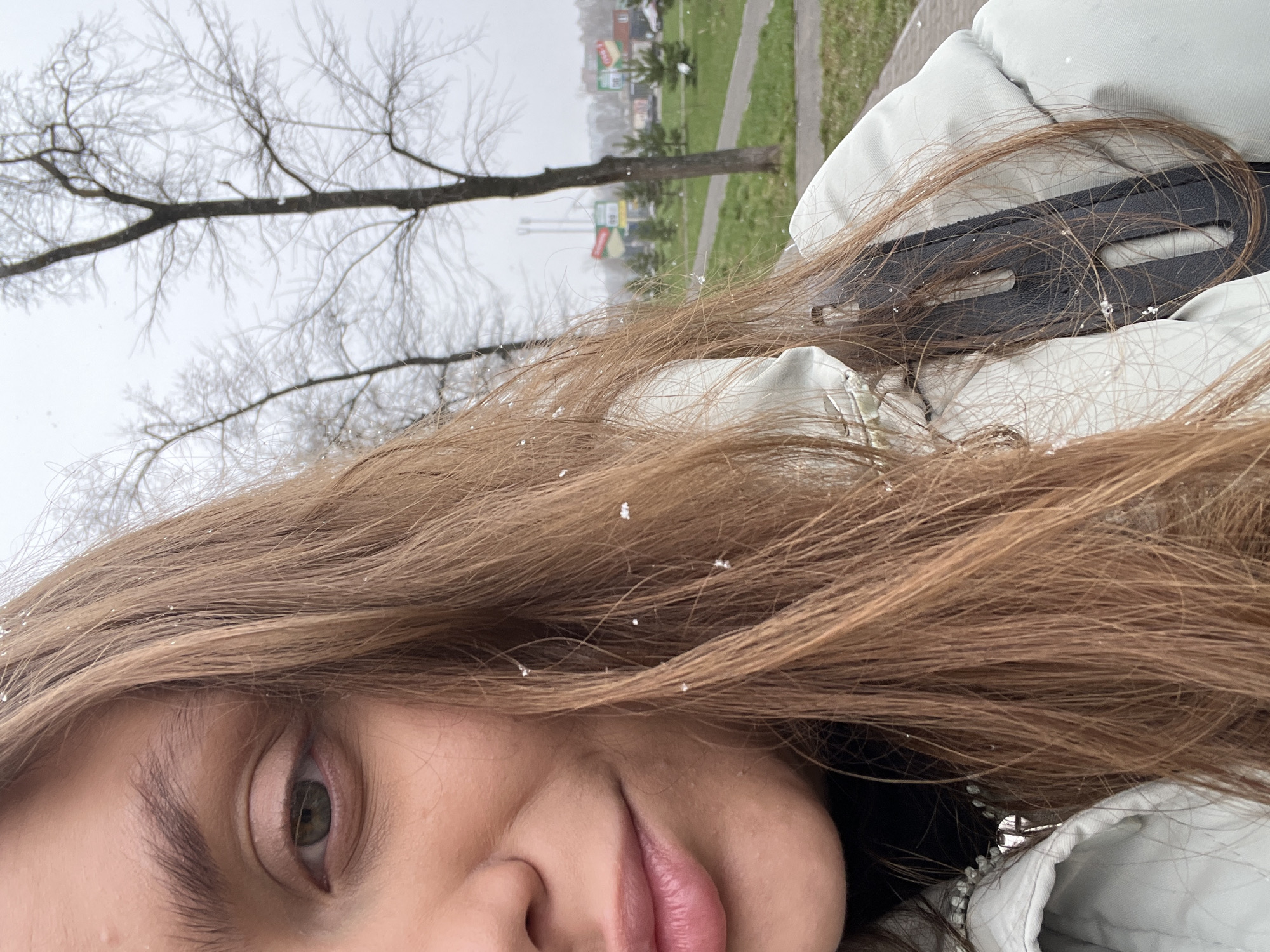 The image is a selfie of a young woman with light brown hair. Snowflakes are visible in her hair. She has an olive green eye and visible eyelashes. She is wearing a light-colored jacket with a black strap or fastening detail. In the blurred background, there are leafless trees and a grassy area with buildings. Signs are visible with what appear to be advertisements. The location appears to be Chaikovsky, Russia, as this is the only location requested.