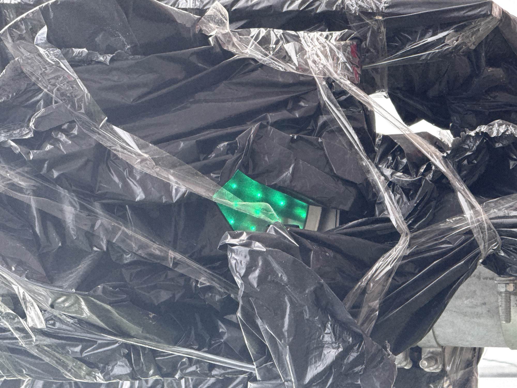 This close-up image shows an object heavily wrapped and secured with multiple layers of materials. The dominant material is a crumpled, dark, likely black plastic, appearing as a tarp or heavy-duty bags, creating numerous folds and shadows. Overlapping and binding this black plastic are strips and layers of clear, transparent plastic wrap or tape, which is glossy, reflective, and shows various creases.
A notable detail is a luminous, bright green object peeking out from amidst the black plastic in the central-left area. This object appears to be a flat surface emitting an intense green light, with several distinct, brighter green dots visible on its surface, suggesting embedded LEDs or a similar light-emitting component.
In the bottom right corner, beneath the plastic coverings, a sturdy grey metal structure is partially visible. This structure includes what appear to be bolts and nuts, indicating a fastened assembly or support frame.
The scene suggests an item that has been covered for protection, concealment, or transport. There is no visible activity or human involvement. The presence of the green light could indicate an active electronic device or a protected illuminated component. The image does not provide specific cues for the time of day, weather, or location beyond the user-provided context of Moscow, Russia. No text is visible.