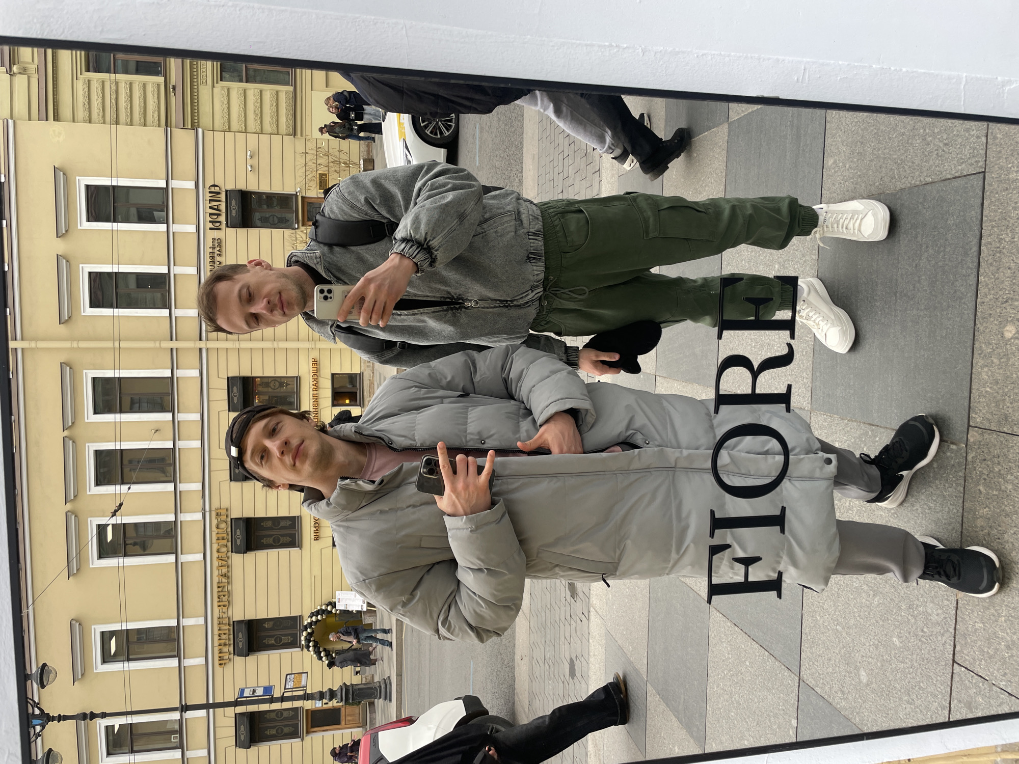 Two men are reflected in a window. The man on the top is wearing a gray jacket, green pants, and white sneakers and holding a white smartphone. The man on the bottom is wearing a gray puffer jacket, gray pants, a black beanie, and black sneakers and holding a black smartphone. The reflections show a building in the background, people walking along the sidewalk, and a parked white vehicle. The letters "FIOR" are visible on the window. The architecture of the building in the reflection is characteristic of the city of Saint Petersburg, Russia.