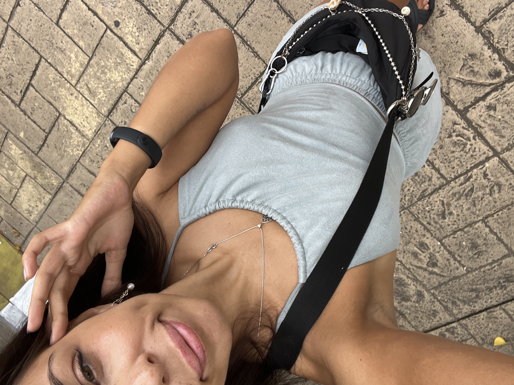 A high-angle selfie captures a woman lying or leaning on patterned grey-brown paved ground. The woman, appearing to be an adult with dark hair, looks up towards the camera with a relaxed and slightly playful expression, her tongue gently visible between her lips.
She is wearing a light blue or greyish-blue sleeveless top with an elasticated neckline. A thin silver necklace with a small pendant (possibly a heart or leaf shape) rests on her chest, with another part of the necklace extending lower. She has a silver earring with a pearl-like drop visible on her right ear, and her left hand is near her head. A black wristband, possibly a fitness tracker, is worn on her left wrist.
A black shoulder bag with silver chain details and silver carabiner-style clips is draped over her, with sunglasses also hanging from one of the clips. A black strap from the bag crosses her body. Her bare arm and part of her leg are visible, along with a foot clad in a black flip-flop or sandal. The paving in the background shows varied textures and joint lines, suggesting an outdoor setting during daytime with bright, even lighting, indicative of warm weather. No visible text is present in the image. The scene conveys a casual and relaxed moment in Kuta Selatan, Indonesia.