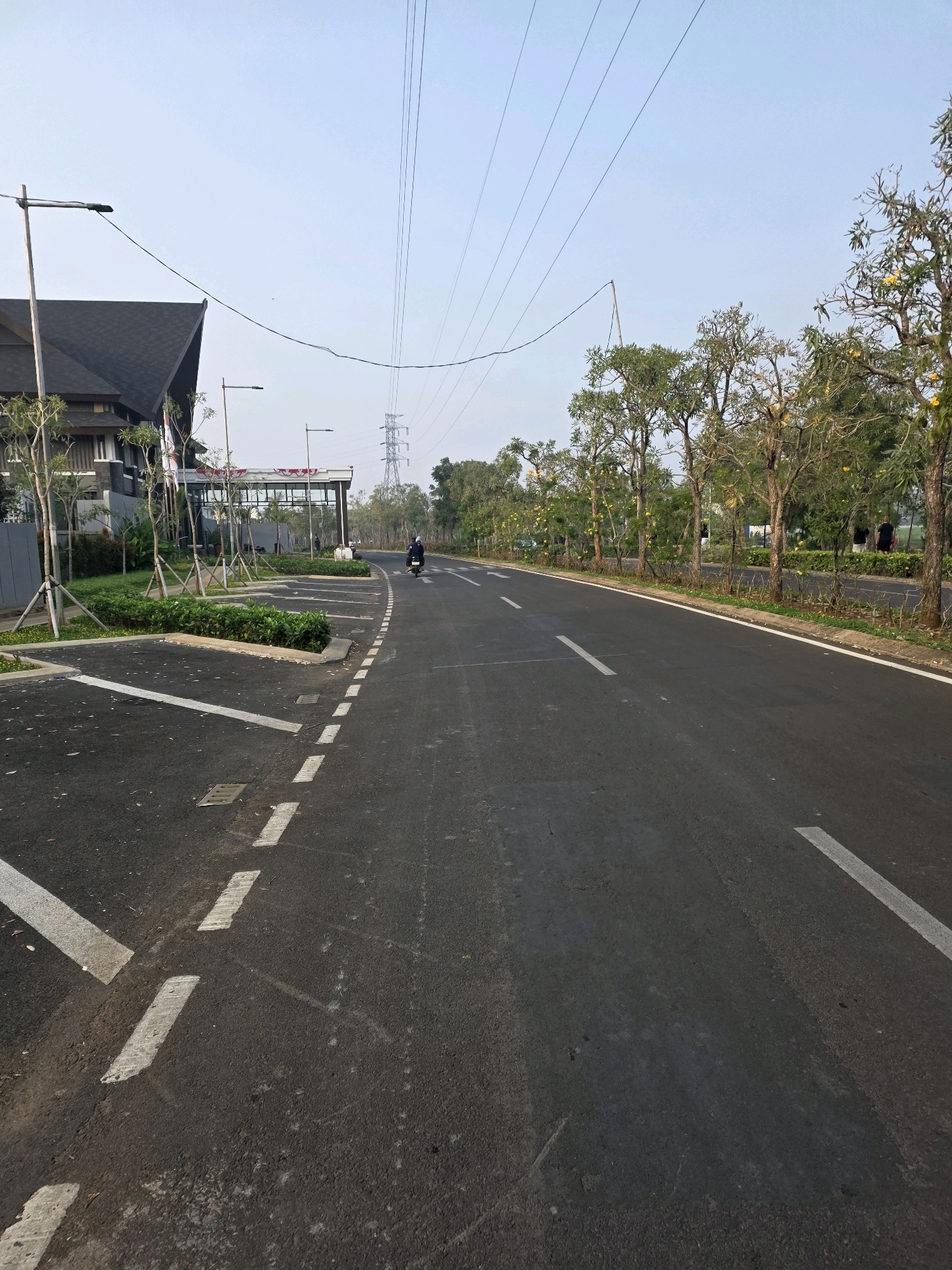 The image depicts a paved road in Bojongsoang, Indonesia, with a parking area to the left. The road features white dashed lines indicating lanes. On the left side of the road, there is a building with a dark roof and several young trees planted in a landscaped area. Further down the road, a motorcycle with one rider is visible. To the right of the road, a line of trees with yellow flowers forms a natural barrier. Overhead, power lines stretch across the sky, supported by tall transmission towers in the distance. The sky is clear and pale blue, suggesting it is daytime.