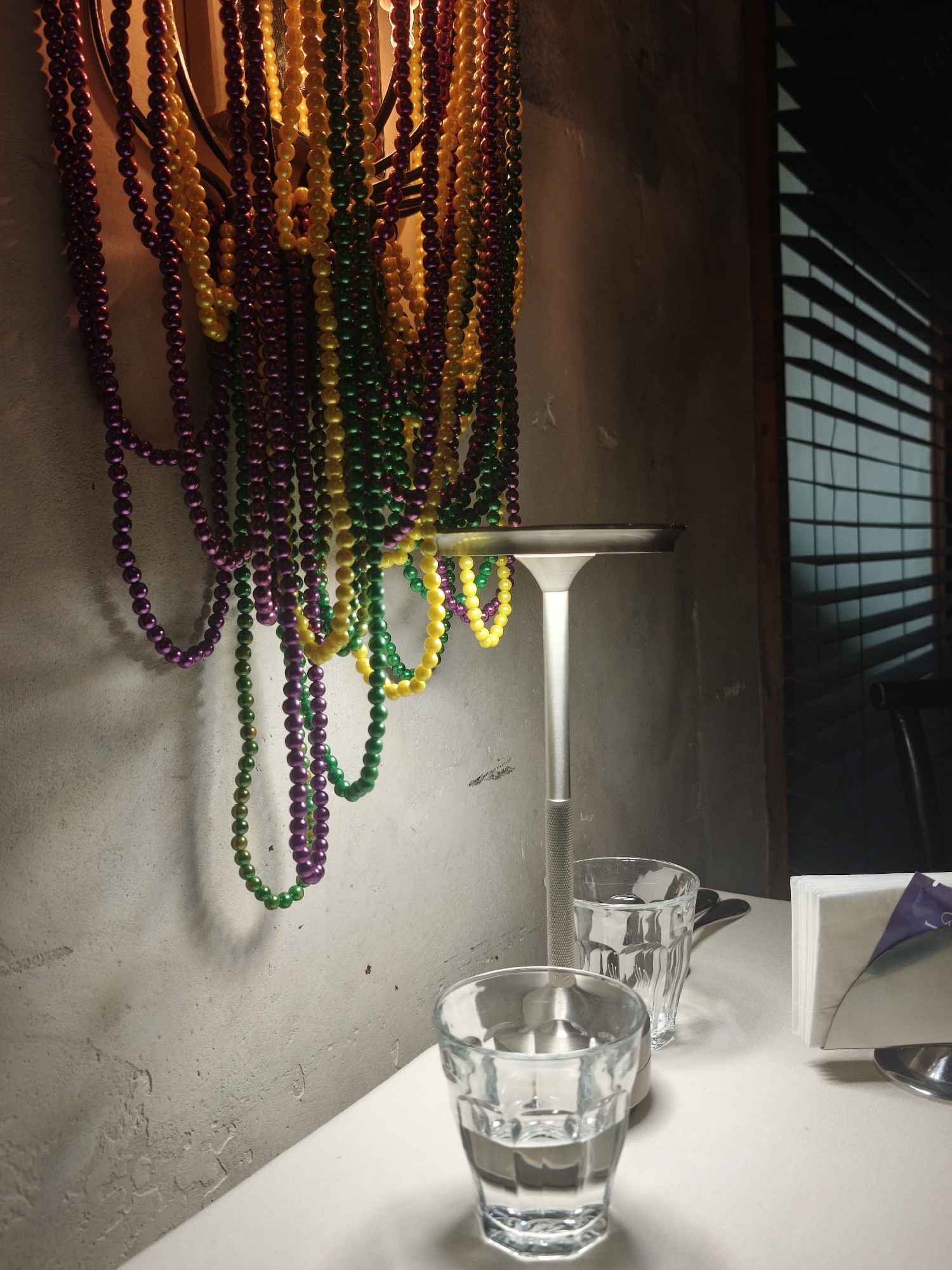 The image depicts a dimly lit indoor scene, likely a restaurant or bar, with a focus on decorative Mardi Gras beads and a modern lamp. The beads, in shades of purple, green, and yellow, are draped in a cluster, suggesting a festive atmosphere. A sleek, silver lamp with a flat, illuminated top stands on a white tablecloth, casting a soft glow. In the foreground, two clear glasses filled with water sit on the table. One glass is closer to the viewer, while the other is slightly behind and to the right. To the right of the glasses, a metal napkin holder filled with white napkins is visible, with a small purple packet peeking out. In the background, a set of horizontal blinds casts a patterned shadow on a textured wall, hinting at a window or partition. The overall mood is somewhat intimate and perhaps a bit mysterious due to the low lighting and shadows. There is no visible text in the image. The setting appears to be Saint Petersburg, Russia.