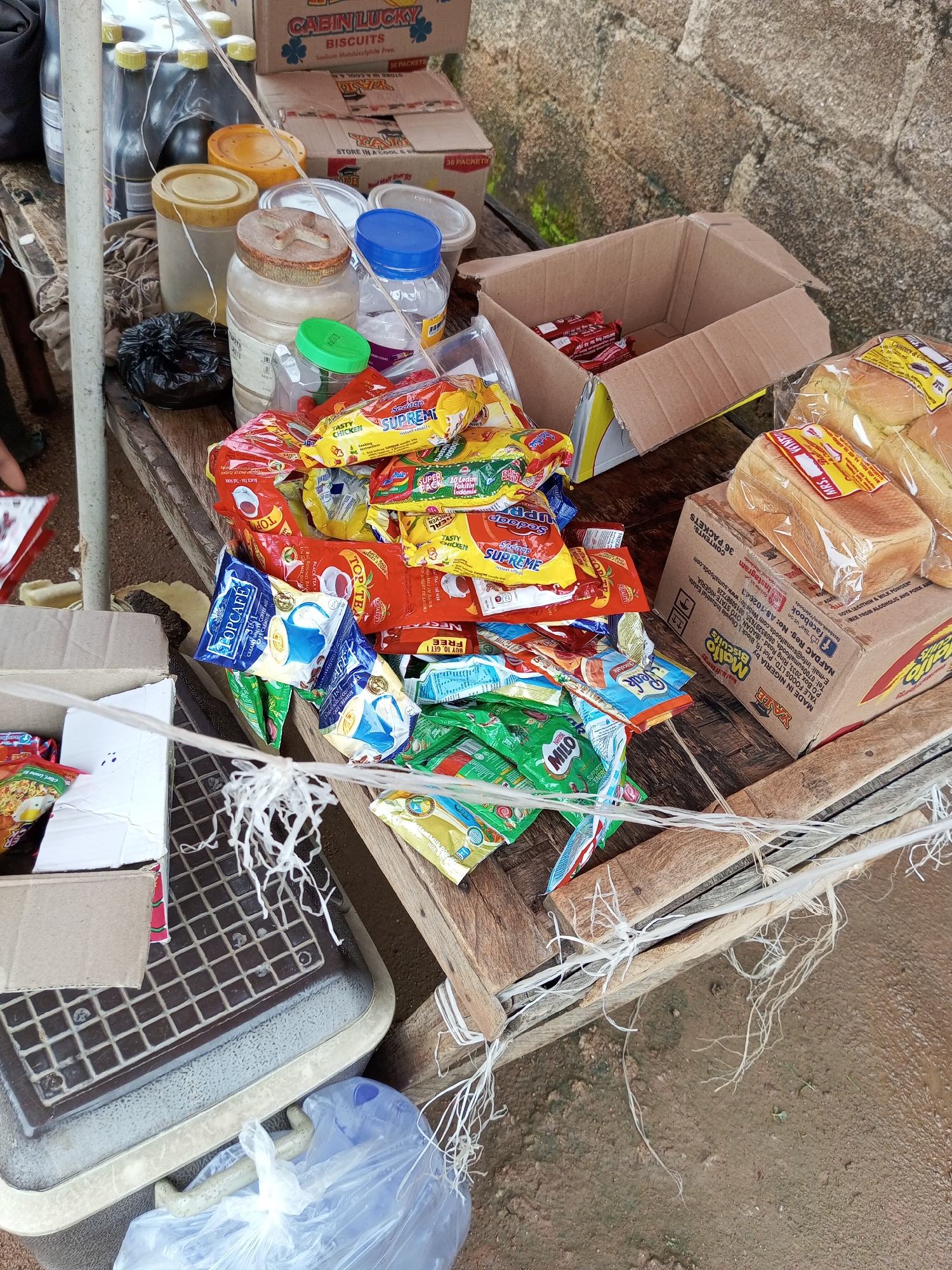 The image depicts an outdoor scene in Bukuru, Nigeria, with various food items and containers laid out on a wooden surface. In the foreground, a pile of individually packaged food items, primarily instant noodles and biscuits, are stacked in colorful wrappers. Some of these include brands like "Supreme," "Tasty Chicken," and "Milo." There are also several plastic jars and containers visible, some filled with yellow and white substances, and one with a blue lid. A large cardboard box, partially open, contains red-wrapped items, possibly more snacks. To the right, two loaves of bread, individually wrapped in plastic, are placed on top of another cardboard box labeled "Mrs." and with Nigerian NAFDAC information. In the background, several dark-colored bottles are lined up, and behind them, a box labeled "Cabin Lucky Biscuits" is visible. The ground appears to be wet and muddy, suggesting it might have rained recently. A white string or rope is draped across the scene, attaching to wooden planks that form part of the display. No people are clearly visible in the image. The setting suggests a small market stall or a personal arrangement for selling or distributing food.