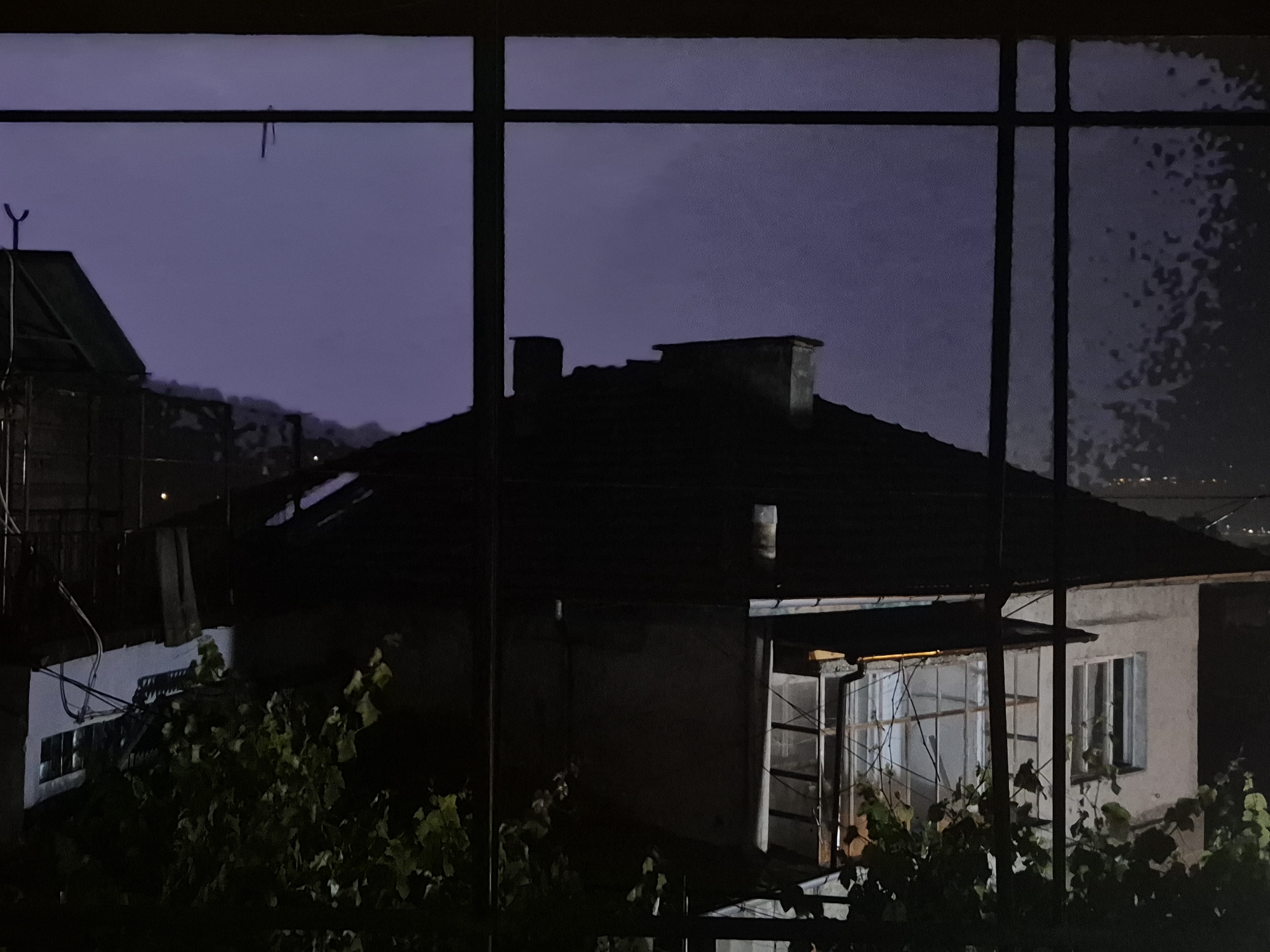 The image captures a nighttime scene viewed through a dark window frame, which divides the composition into several sections. The sky above is a deep, desaturated purple-blue, indicating the late hour, and appears slightly hazy or cloudy.
In the mid-ground, silhouetted against the dark sky, are several houses or buildings. One prominent house in the center has a dark, sloped roof with two distinct chimneys. To the left, another partial building is visible, featuring a Y-shaped antenna or structure on its roof. In the foreground and surrounding the houses, lush green foliage and vines are visible, particularly on the lower left and center-right.
One of the houses in the mid-ground has a window glowing with a pale, warm light, revealing a grid-like structure within, possibly bars or a complex window frame. Adjacent to this, another window appears darker, possibly with closed shutters. Some wires or cables are attached to the side of a white-walled structure on the left. A faint, possibly hanging object is seen suspended from a wire or line against the sky in the upper left section.
Towards the far right horizon, especially visible through the window panes, there are subtle patterns of light, suggesting a distant city or settlement, blurred by distance or atmospheric conditions. The overall lighting is dim, with most elements cast in shadow, emphasizing the artificial light emanating from the single window. There is no visible activity or identifiable text within the scene.